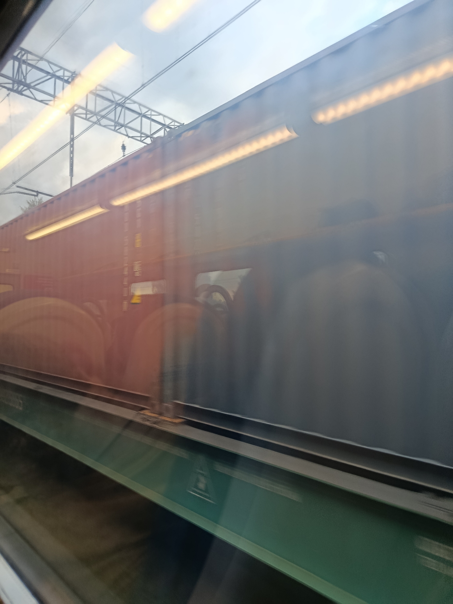 An exterior view from a moving train in Elektrougli, Russia, captures a long, multi-unit cargo train. The train cars are primarily a reddish-brown hue, with some appearing darker and blurred due to motion. Bright, elongated yellow lights run along the upper portion of the train cars, suggesting interior illumination or external lighting. The train is on a track, with dark, metallic rails visible at the bottom of the frame. Overhead, power lines and support structures are visible against a cloudy, overcast sky. The image is distorted by a reflective surface, likely the train's window, creating streaks of light and a slightly hazy appearance. The overall scene depicts transportation infrastructure and cargo movement in an urban or semi-urban environment.