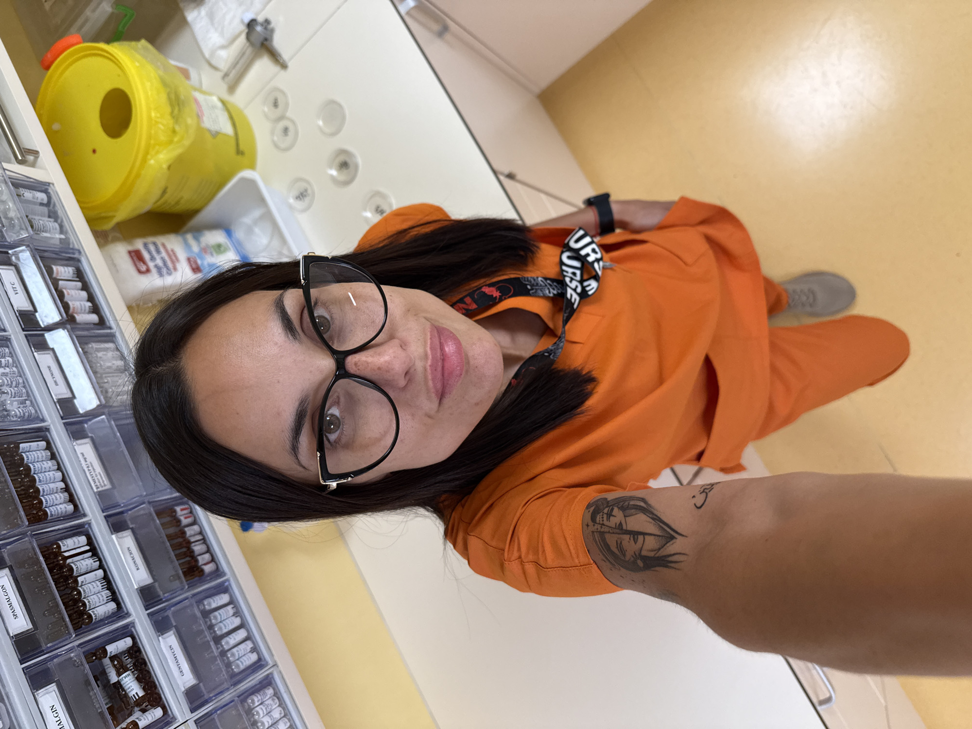 A woman in an orange uniform and glasses is taking a selfie in what appears to be a medical setting. She has dark hair and a tattoo on her forearm. She is standing near drawers filled with vials labeled with different medications, including SPASMALGON, GENTAMYCIN, VIT C, KONAKION, and others. There's a yellow sharps container and medical supplies on the countertop. The floor is a light yellow. The city is Pleven, Bulgaria.