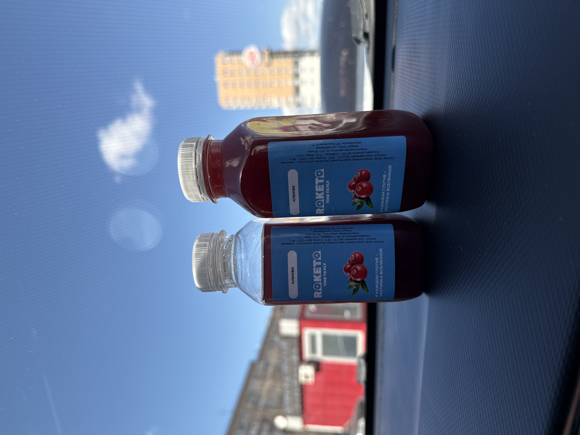 The image shows two bottles of "Roketo" cranberry juice. The label on the bottle reads "Roketo Time to Fly" with an image of cranberries. The text is in Russian. The juice is a deep red color. The bottles are sitting on a dark, textured surface, likely the dashboard of a car. Outside the car window, there is a view of Izhevsk, Russia. A large yellow building with a red emblem on top is visible in the background. The sky is blue with scattered clouds. In the lower part of the image, another building with red and white exterior is visible.