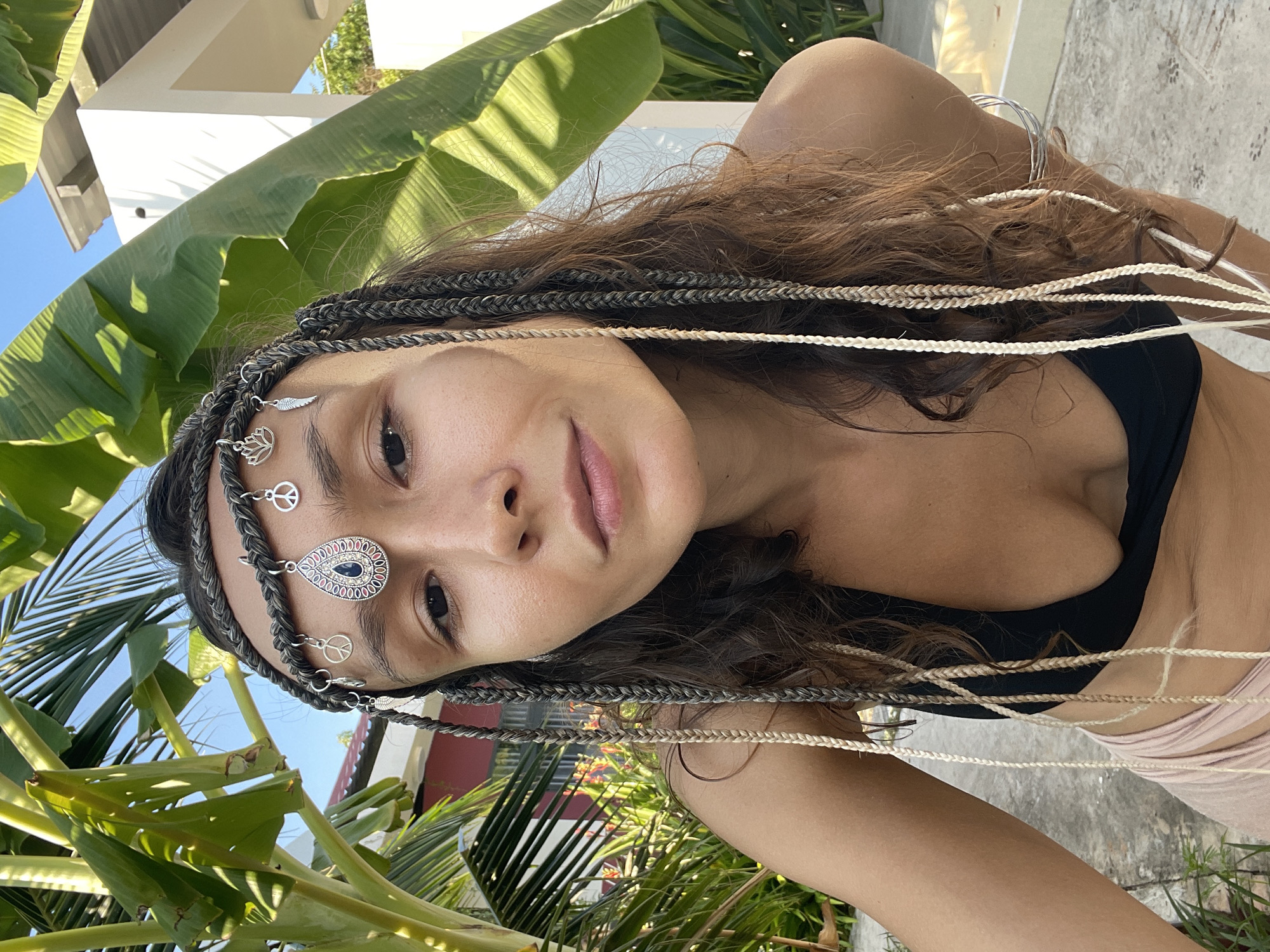 A woman is photographed from the chest up in an outdoor setting. She is wearing a black bikini top and a beaded headband adorned with peace signs and a teardrop-shaped pendant with a blue stone. Her hair is styled in numerous braids, some of which are mixed with lighter colored strands. She is looking directly at the camera with a neutral expression. In the background, lush green foliage, including large palm leaves, is visible. A white building with a sloped roof and a section of a red building are also present. The scene appears to be a sunny day. The location is identified as Mũi Né, Vietnam.