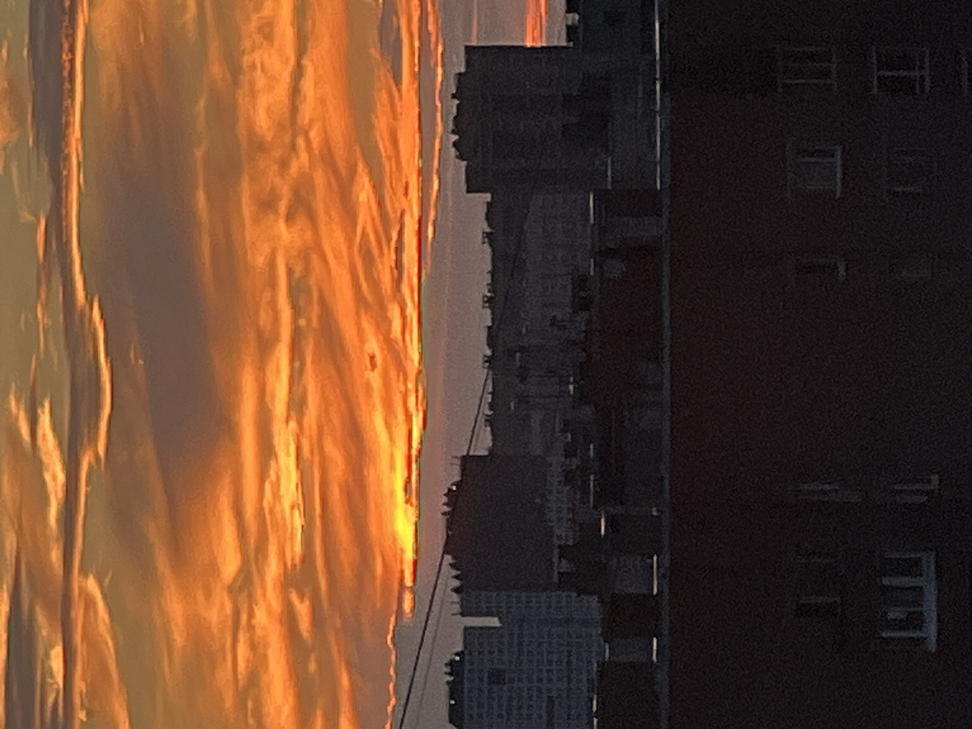A dramatic sunset illuminates the sky over the city of Sinyavino, Russia. The sky is a vibrant display of orange and yellow hues, with streaks of pink and purple creating a textured, layered effect in the clouds. Silhouetted against this fiery backdrop are the indistinct shapes of urban buildings. In the foreground, dark, blocky structures with visible window outlines dominate the lower portion of the frame, suggesting a residential or industrial area. The overall scene captures a moment of natural beauty as the day ends, casting a dramatic glow over the cityscape.