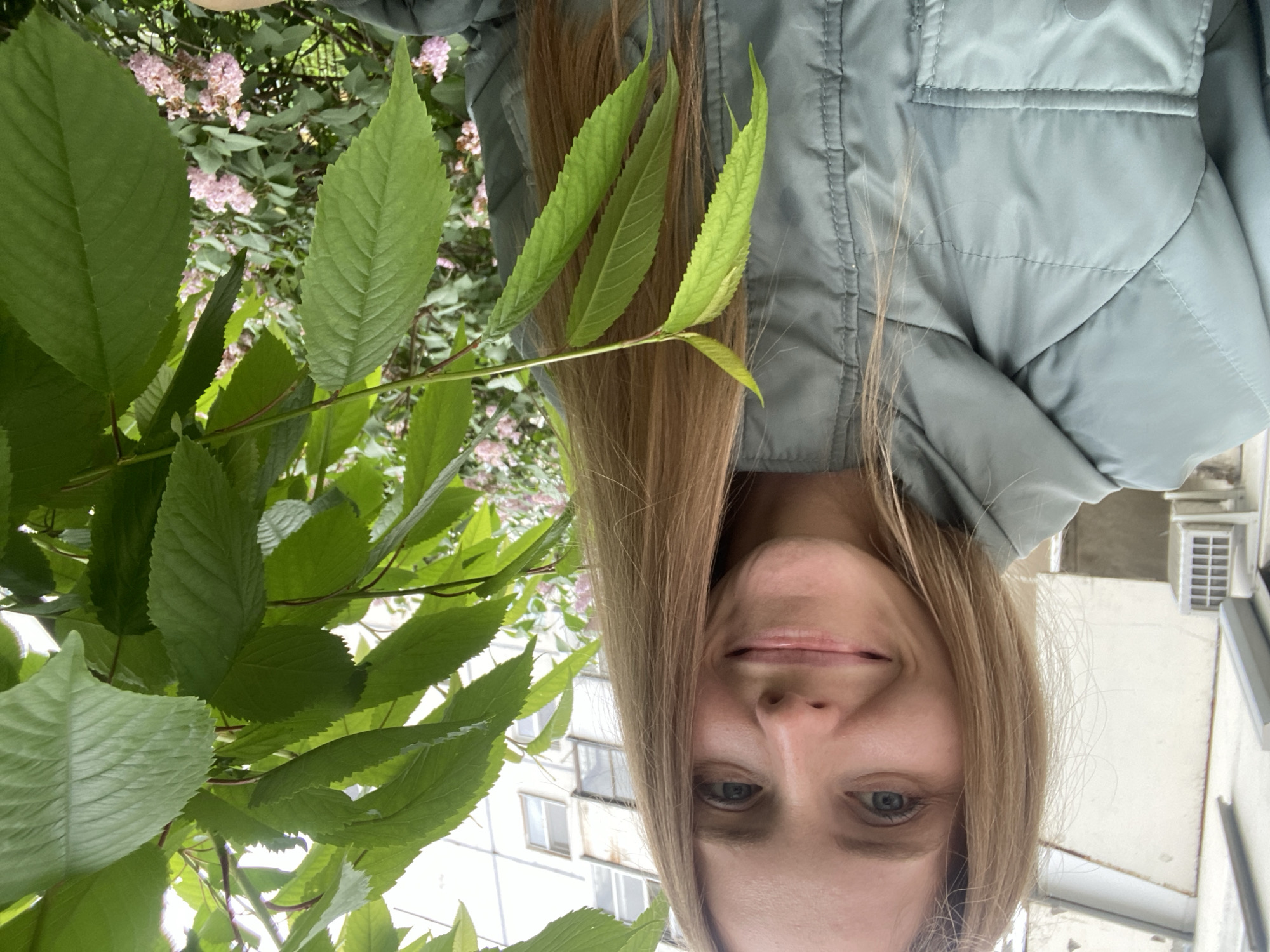 A young woman with long blonde hair is captured in a close-up, outdoor, selfie-style photograph. She is smiling directly at the camera, her face framed by her hair and some green leaves on the right side of the image. She is wearing a light green, puffy jacket with visible stitching and a pocket. In the background, a building with windows and an air conditioning unit is partially visible, along with a bush with small pink flowers. The setting appears to be an urban environment.
