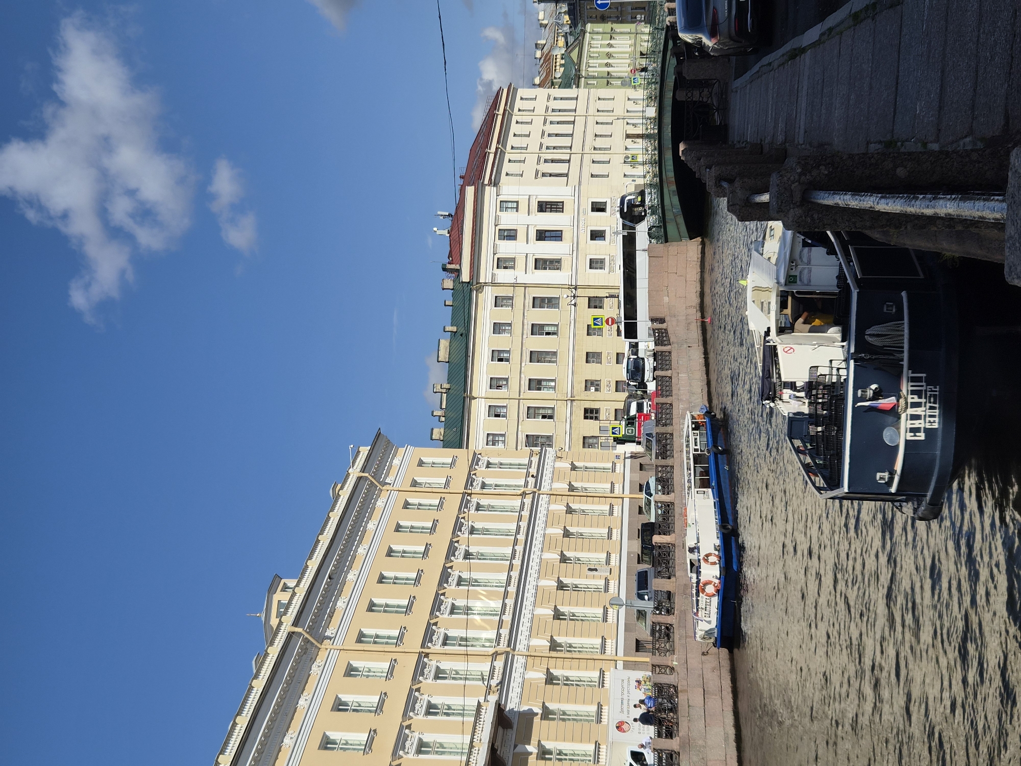 This is a vertical, eye-level, medium shot of a sunny day along a canal in a city. On the left, a long, pale yellow building with multiple windows and ornate white trim runs the length of the canal. A dark grey stone embankment separates the canal from the building, with a black metal railing running along the top. Several people are visible on the embankment, standing and walking. A white delivery truck with Russian text and images is parked near the embankment. Several cars are parked behind the embankment.
A light blue and white boat with orange life preservers is docked in the canal. Further down the canal, a dark blue boat with a white cabin and the Russian flag is also docked. A green bridge with an arched span crosses the canal in the background. Beyond the bridge, more buildings line the canal, one with a red roof and another with a green roof. The water of the canal is dark and choppy, reflecting the buildings and sky. The sky is a bright blue with a few scattered white clouds.
On the right side of the image, a paved walkway runs alongside the canal, bordered by a dark stone embankment with a rough-hewn stone railing. A dark-colored car is parked on the walkway. The scene appears to be a picturesque urban setting, possibly a European city known for its canals.