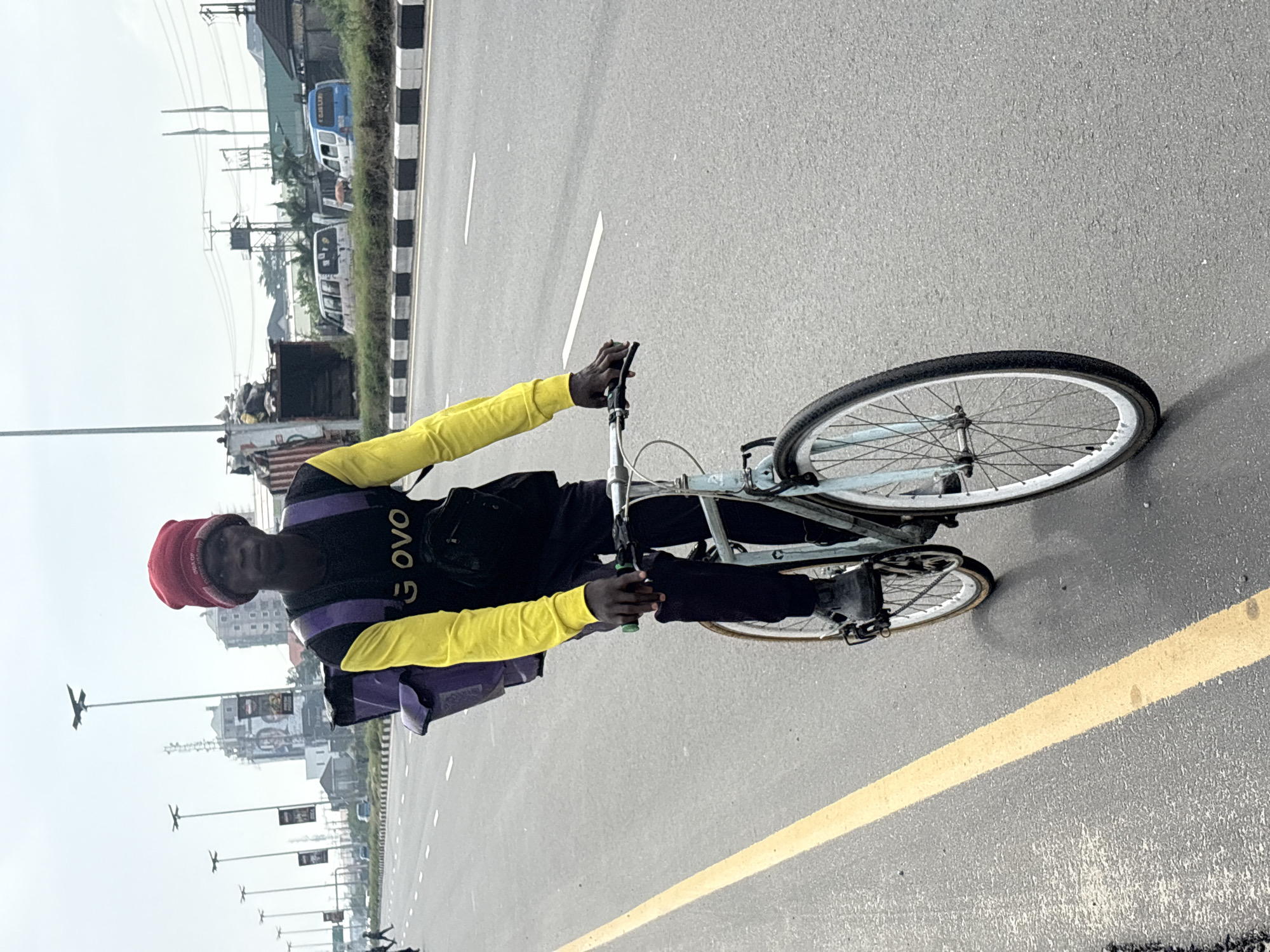 A man with dark skin and a red beanie is riding a light blue bicycle on a paved road in Igboefon, Nigeria. He wears a black shirt with the word "GOVO" visible, a purple vest with yellow sleeves, and black pants. He is looking towards the camera. The road has yellow lines marking lanes. In the background are buildings, streetlights, power lines, and vehicles. The sky is overcast.