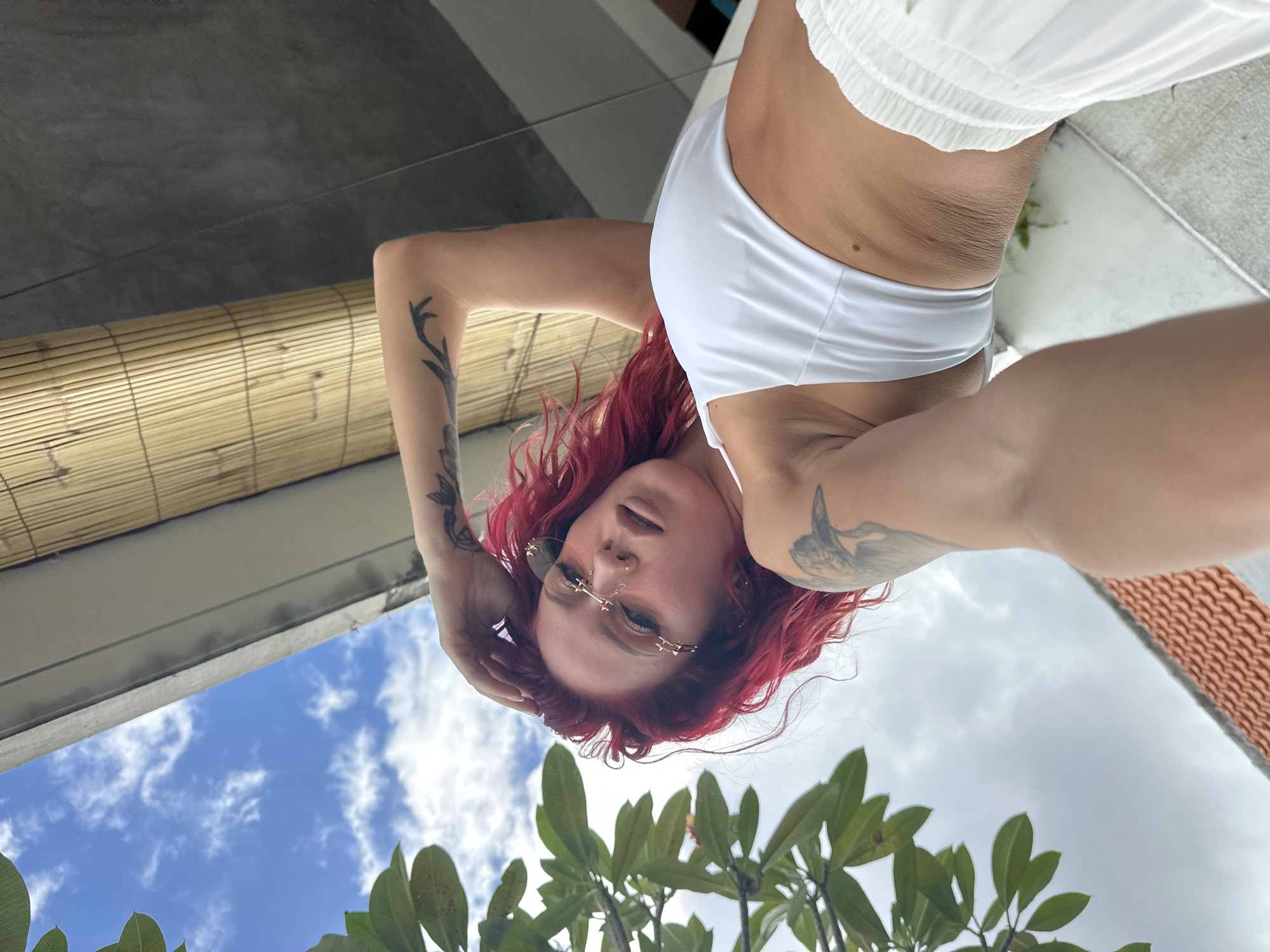 A woman with red hair is pictured from a low angle. She wears a white bikini top and white shorts. She has multiple tattoos. The background includes a blue sky with clouds, green foliage, a bamboo shade, and a concrete structure. The location is implied to be in North Kuta, Indonesia.