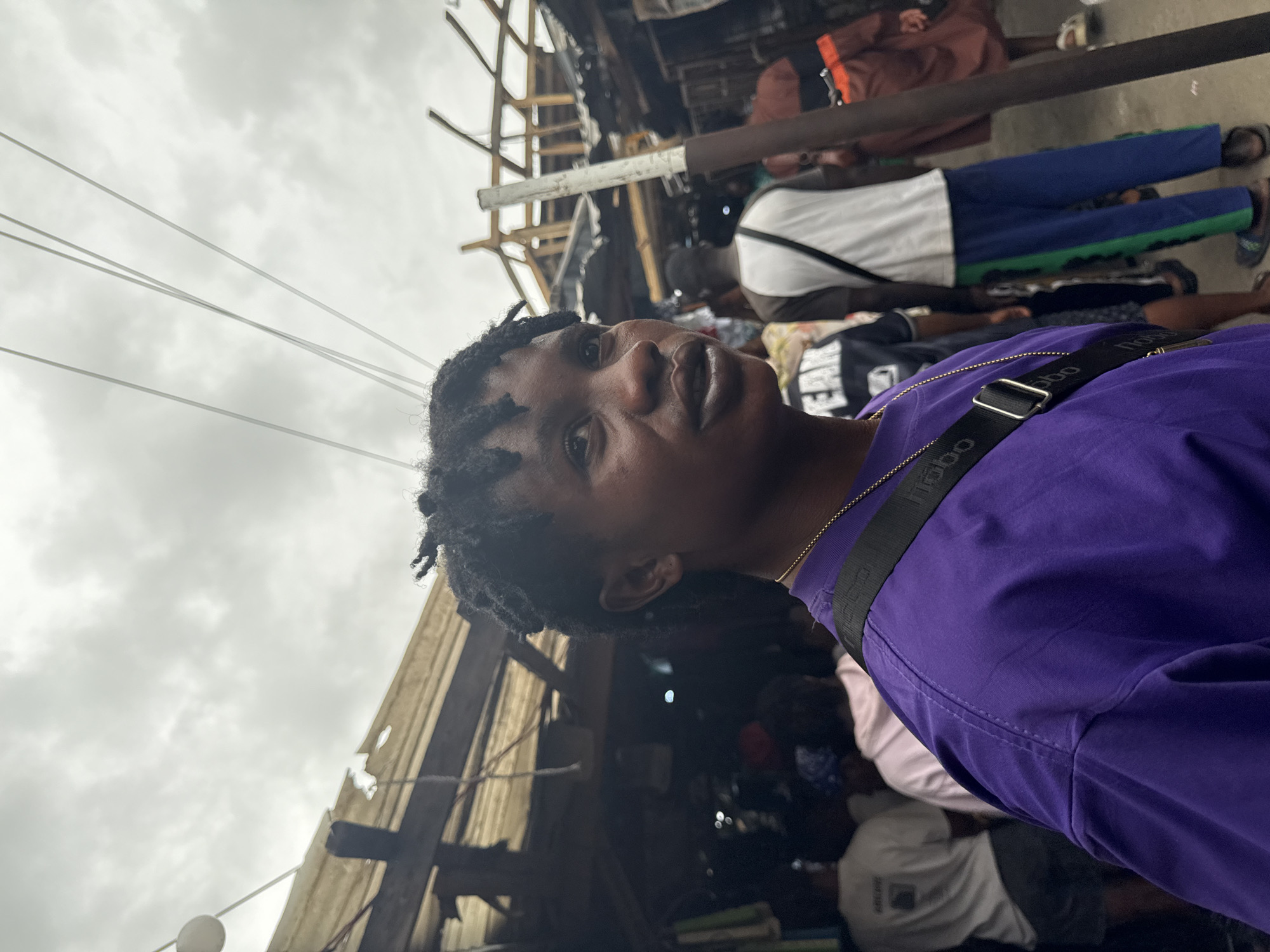 A young person with dreadlocks, wearing a purple t-shirt and a gold chain, is the subject of this medium shot portrait. They are looking off to the right, with their mouth slightly open. Behind them, a crowded outdoor market scene unfolds. Several other people are visible, some wearing t-shirts and casual wear. The background is filled with market stalls and overhead structures, appearing somewhat dilapidated and possibly damaged, with exposed wood and corrugated metal. The sky above is overcast and cloudy. The overall impression is of a bustling, informal marketplace, likely in a developing urban area. The context provided indicates this scene is in Lekki, Nigeria.