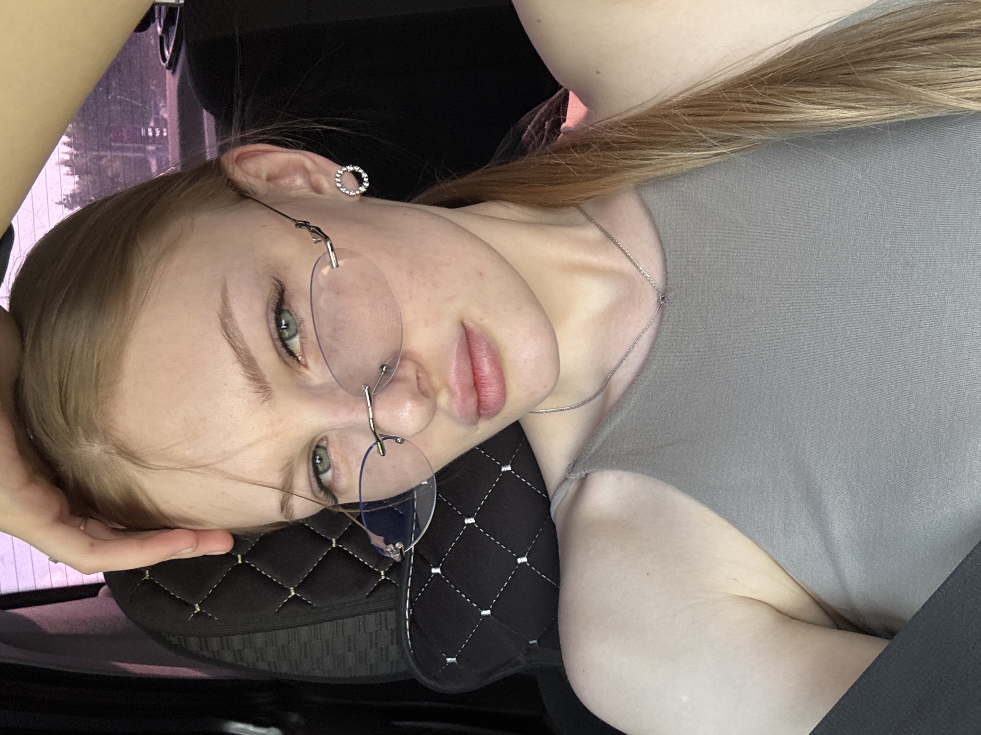 A young woman with blonde hair and fair skin is in a car. She is wearing glasses, a gray shirt, and a small circular earring in her left ear. She also has a delicate silver necklace. The woman is looking at the camera with her head tilted to the left, resting on a black quilted headrest. Her left hand is positioned near her forehead. The car's interior is visible, including the black seat and a glimpse of the outside through the window. The location is not definitively discernible from the image, nor the city of Perm.