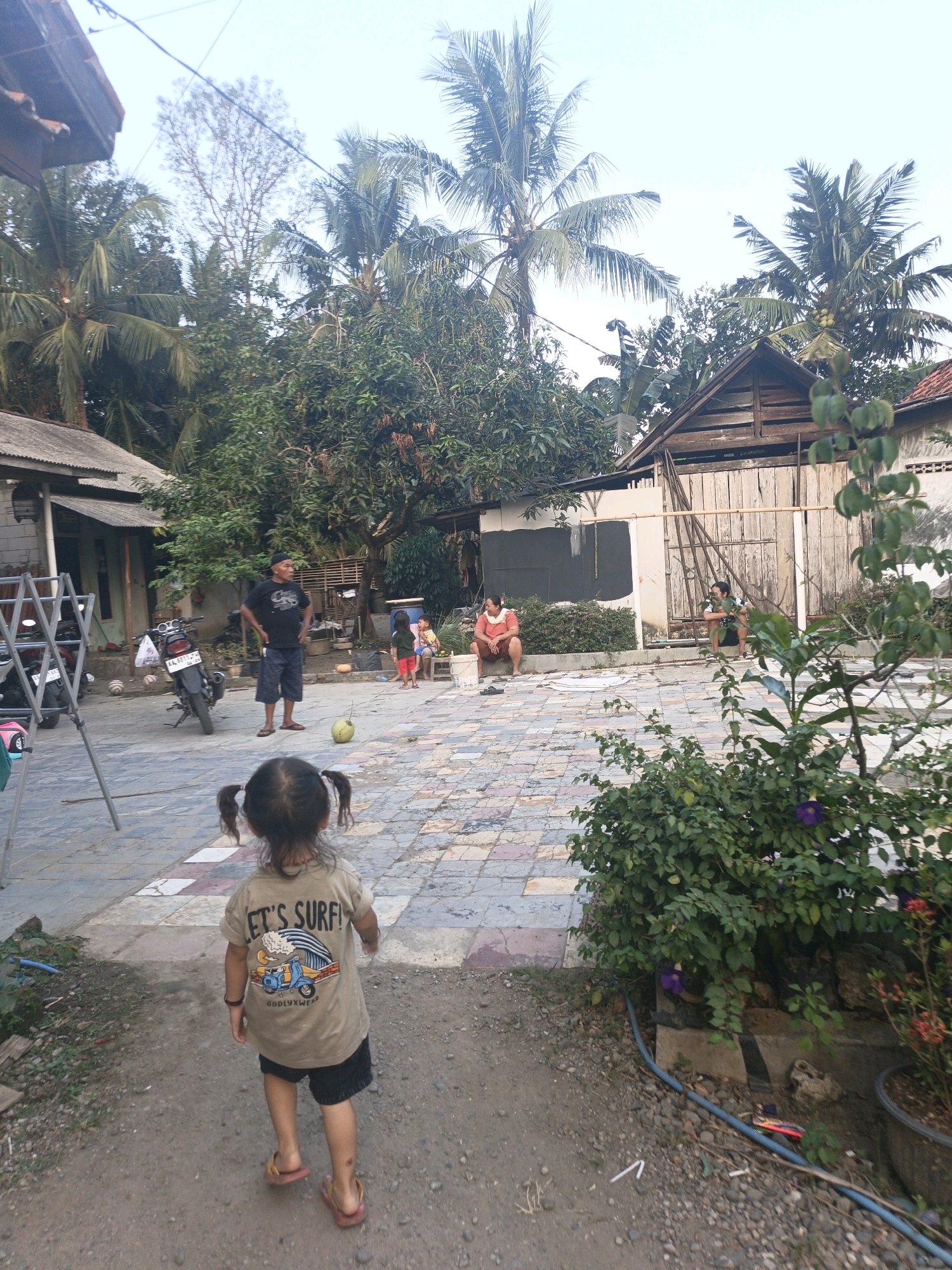 This is a vertical, eye-level, medium shot of a residential courtyard in Kamulyan, Indonesia. In the foreground, a young girl with pigtails walks away from the camera. She is wearing a tan t-shirt with a graphic that reads "LET'S SURF!" and black shorts. Her feet are bare except for flip-flops. Behind her, a paved courtyard is visible, with a few scattered coconuts and a soccer ball. To the left, a motorcycle is parked next to a building with a gray roof and white walls. In the middle ground, several adults and children are present. A man in a black t-shirt and shorts stands with his hands on his hips, looking to the side. A woman in an orange shirt is sitting on a ledge, and a few children are playing. In the background, a wooden structure with a pitched roof is visible, along with lush green trees and palm trees. The sky is overcast.
