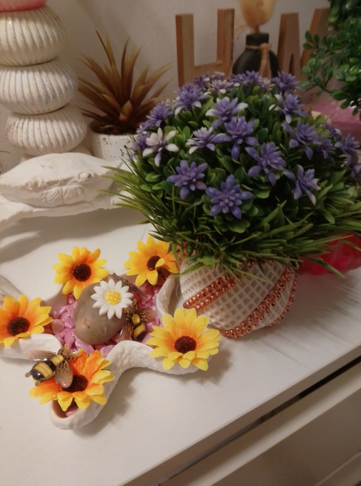 A decorative arrangement is displayed on a white surface. In the foreground, several yellow, artificial sunflowers with dark brown centers are placed around a white, sculptural element resembling a crab. Two small, decorative bees are positioned on two of the sunflowers, and another bee is near a small, grey stone with a white daisy-like embellishment. Behind the sunflowers, a potted artificial plant with numerous small, purple flowers and green foliage sits in a decorative white pot. To the left of the sunflowers, a stack of three white, ribbed decorative objects is visible. In the background, wooden letters spelling "HOM" are partially visible, along with a dark bottle and some greenery. The scene appears to be an indoor display, possibly in a home in Wedel, Germany.