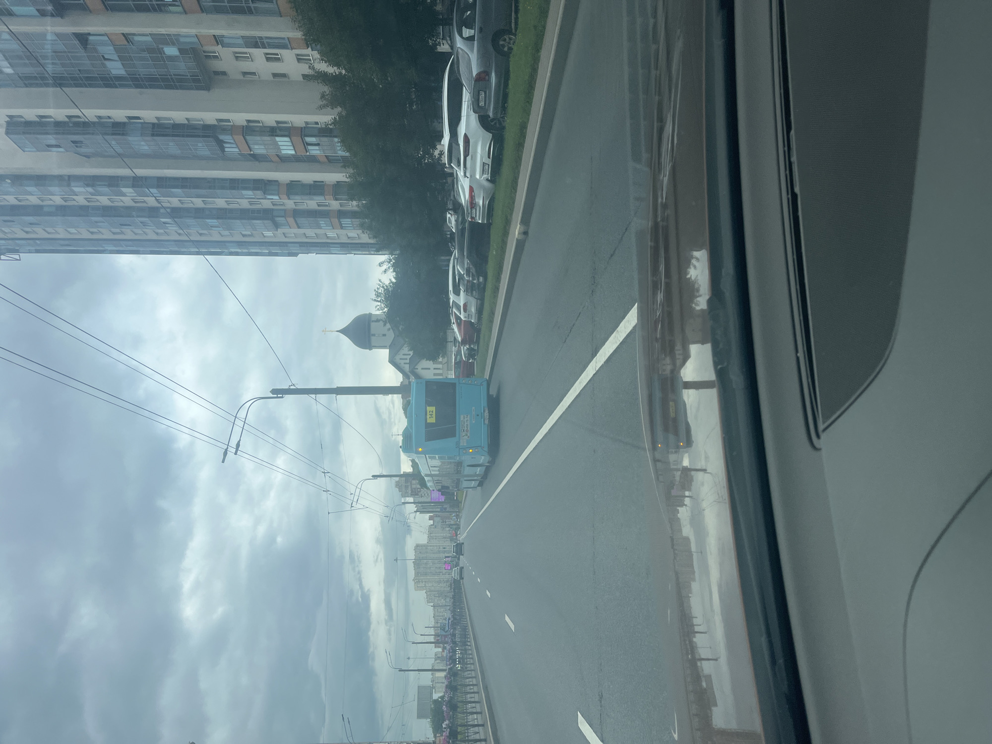 The image shows a view from inside a vehicle looking out at a street scene in Saint Petersburg, Russia. A blue bus with the number "142" displayed is driving away from the camera in the right lane of a multi-lane road. To the right of the road, there is a row of parked cars and trees, behind which stands a tall, modern residential building. To the left of the road, there is a sidewalk with flower planters and more residential buildings in the distance. Overhead, there are trolleybus wires and streetlights. The sky is overcast with a lot of clouds. The overall scene depicts a typical urban environment on a cloudy day.