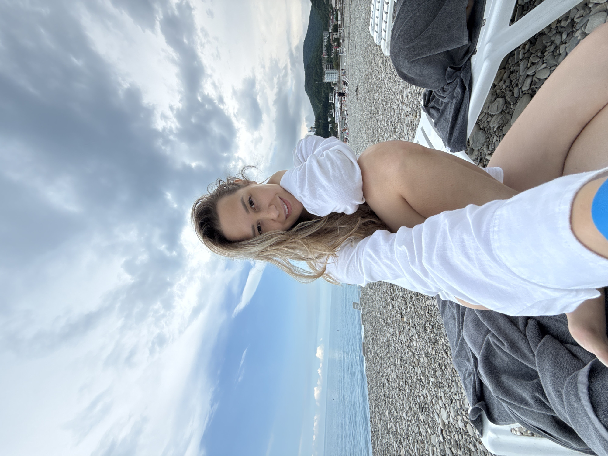 A woman is sitting on a beach chair on a pebble beach, facing the camera. She is wearing a white long-sleeved shirt. The sea is to her left, and a cloudy sky is above. In the background, there are more beach chairs, some people, and buildings nestled in the hills. The location appears to be Nebug, Russia, as indicated by the context.
