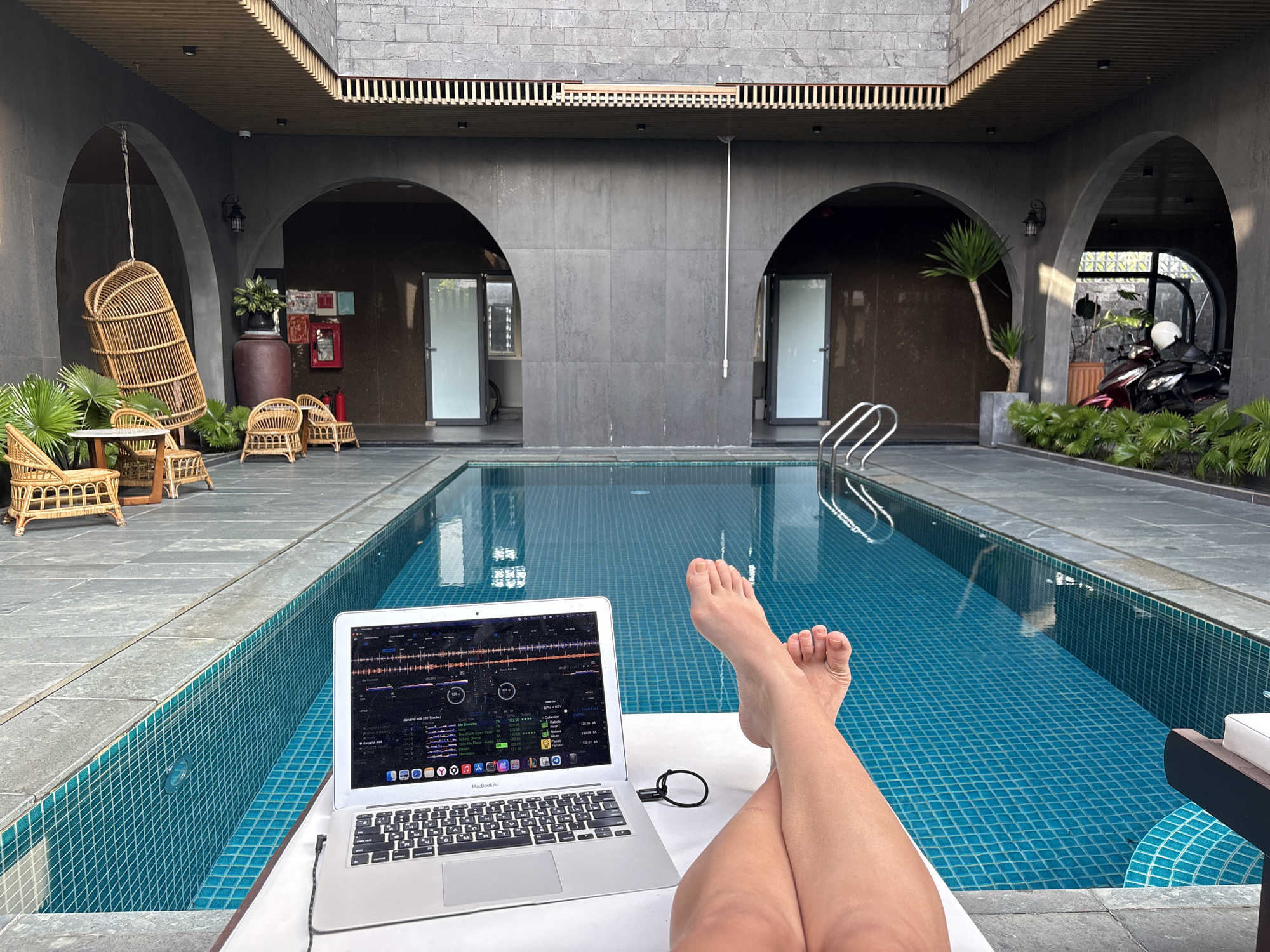 A medium indoor shot shows the lower legs and feet of a person reclining in a lounge chair in the foreground, with a laptop resting on the chair. The screen displays a DJ interface with musical selections. To the side of the laptop is a black charging cable. The person's legs are crossed, with bare feet pointing towards the laptop.
Behind the lounger is a pool of water with a turquoise tiled bottom and sides. Beyond the pool is a covered walkway with arches. On the left side of the walkway are rattan chairs, a hanging rattan chair, and a small table. The walkway is paved with grey stone tiles.
On the right side of the walkway is a motorcycle parked near foliage. The building has a grey facade with decorative latticework near the roof. The image was likely taken in a resort or villa in the city of Đà Nẵng, Vietnam.