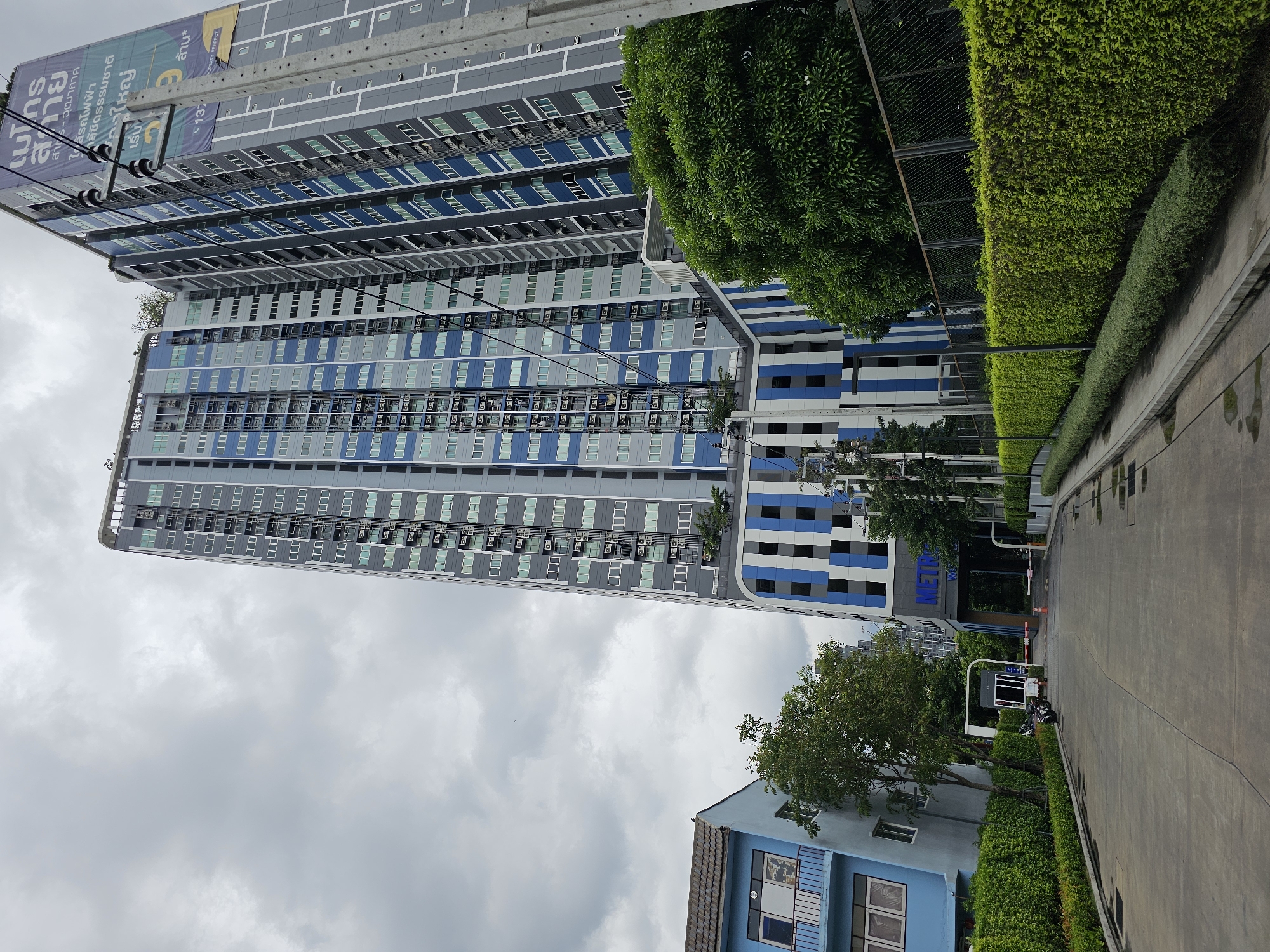 The image depicts the exterior of a modern condominium building in Bangkok, Thailand, on a cloudy day. The primary subject is a tall, multi-story residential building with a facade characterized by alternating sections of gray and blue vertical stripes, and numerous windows with light-colored frames. Adjacent to this building, to the left, is a smaller, light blue building with a tiled roof and windows. A well-maintained green hedge lines the foreground, separating a paved road from the buildings. A security gate with barriers is visible at the entrance to the condominium complex. Electrical poles with visible wiring stand in front of the main building. The sky is overcast with a significant amount of clouds, suggesting a humid or rainy climate typical of Bangkok. The scene conveys a sense of urban living with a focus on residential architecture and landscaping.