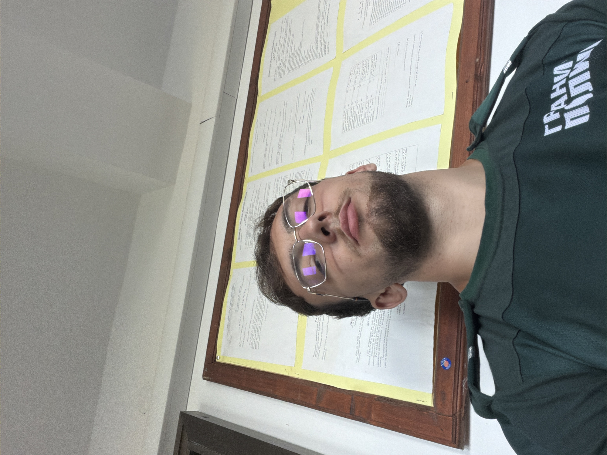 A young man with glasses and a beard looks up at the camera. He is wearing a dark green shirt with white text that reads "ГРАНИЧНА ПОЛИЦИЯ" which translates to "BORDER POLICE". Behind him is a bulletin board with several documents pinned to it. The ceiling is visible above the bulletin board. The image appears to have been taken indoors, possibly in a government building or office. The style of the bulletin board and the surrounding architecture suggest a location in Bulgaria, specifically Rezovo, a town on the Black Sea coast known for its border with Turkey.