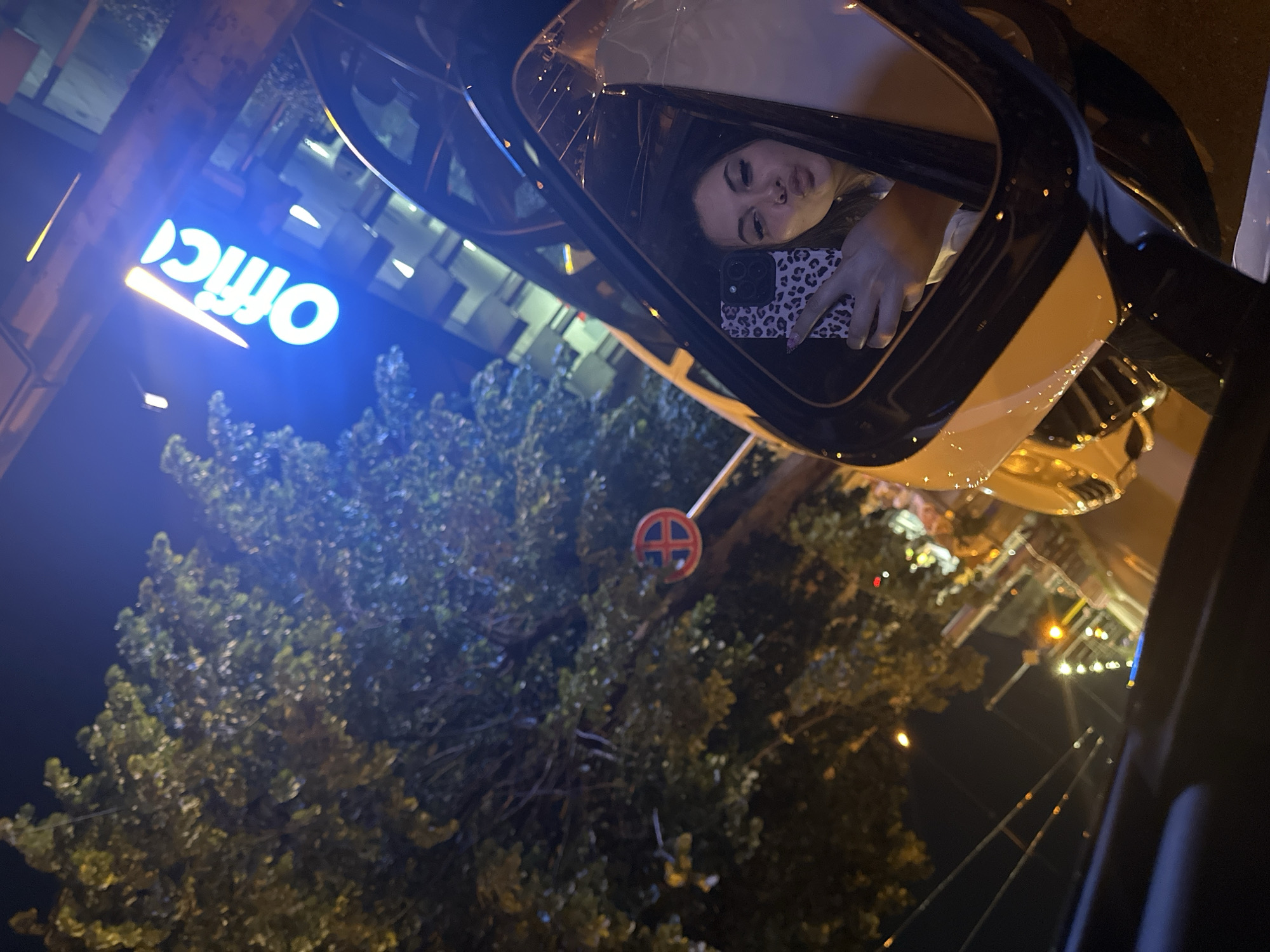 A low-angle, long shot is taken at night, showing a woman inside a car. The woman is holding a phone with a leopard print case and has her lips pursed. Reflected in the car window is a lit-up "Office" sign, a tree, and a street sign. The surroundings appear to be an urban area at night.