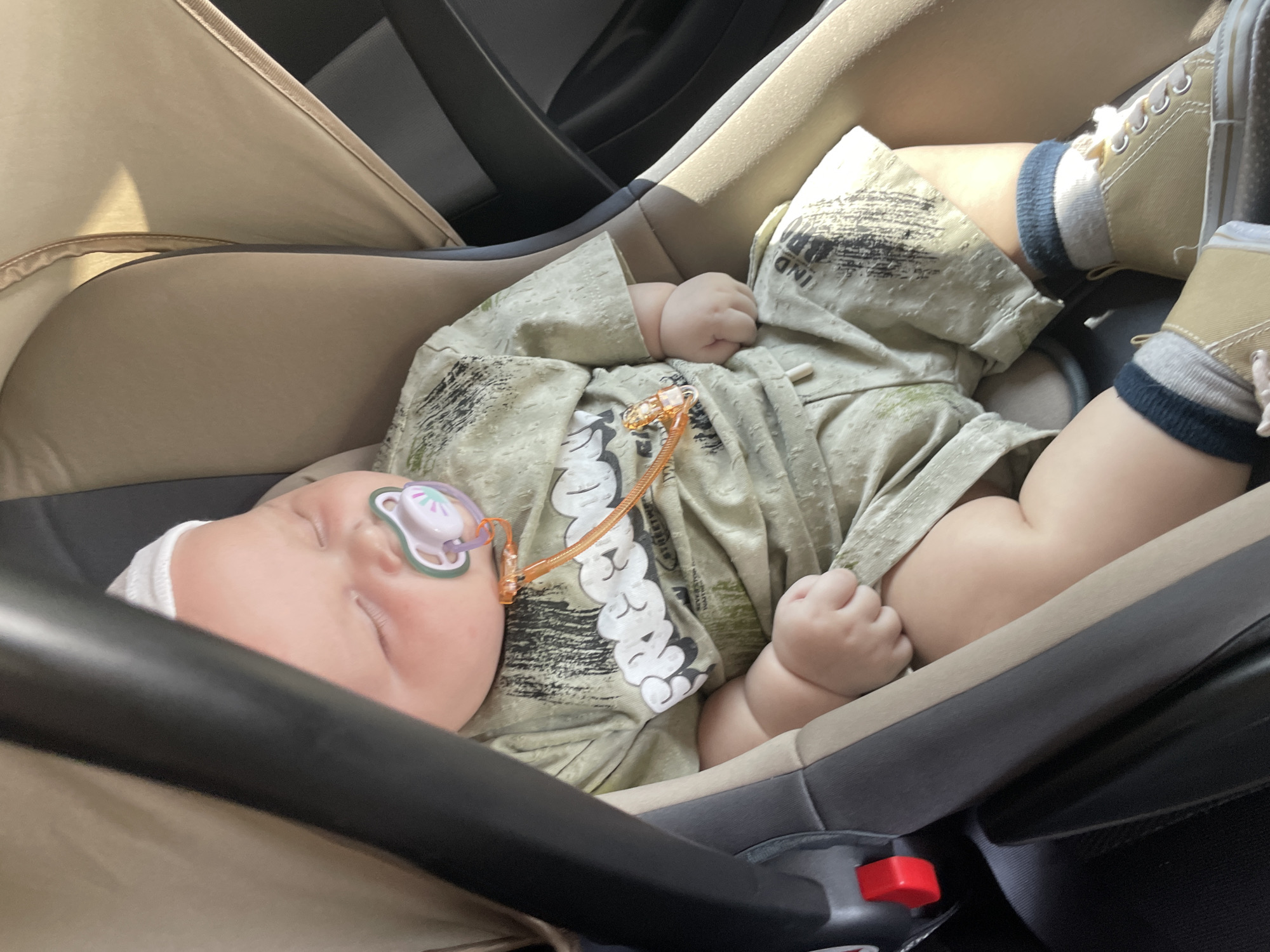 The image shows a baby sleeping in a car seat. The baby is wearing a light green patterned outfit and a white cap. A pacifier is in the baby's mouth. The car seat is tan and grey, with a black harness. The scene appears to be inside a car, with sunlight filtering in from the side, suggesting it is daytime. The overall mood is peaceful and calm, as the baby is asleep. No text is visible in the image.