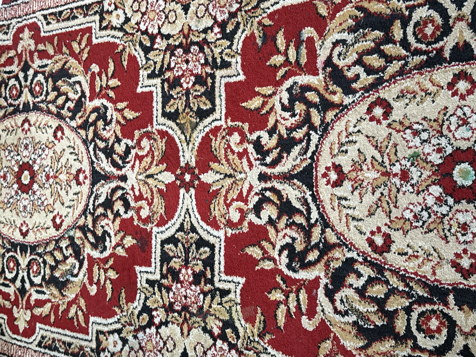 The image shows a close-up view of a red, black, and cream-colored carpet with an intricate floral and paisley pattern. The carpet appears to be made of synthetic fibers and shows some signs of wear and tear, including a few small dark stains and some frayed edges. The pattern is repetitive and symmetrical, with medallion-like designs and scrolling floral motifs. The colors are vibrant, though the lighting makes it difficult to determine the exact shade of red and cream. The perspective is from above, looking down at the carpet. The image does not provide any information about the location in Votkinsk, Russia, or any people or actions.