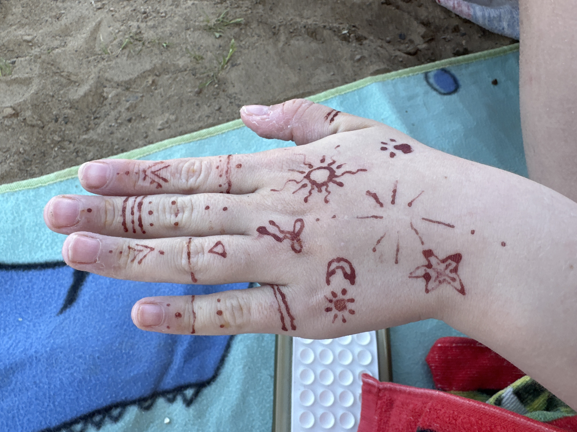A fair-skinned hand is displayed against a backdrop of a blue patterned towel, sand, and parts of other objects. The hand has multiple brown-red temporary tattoos or markings. The tattoos include geometric shapes such as triangles, lines, and arrows on the fingers. There are images of stars, suns, a paw print, and other abstract designs on the back of the hand. The thumb appears irritated. The environment suggests an outdoor setting, likely a beach or sandy area. The location is not determinable from this image.