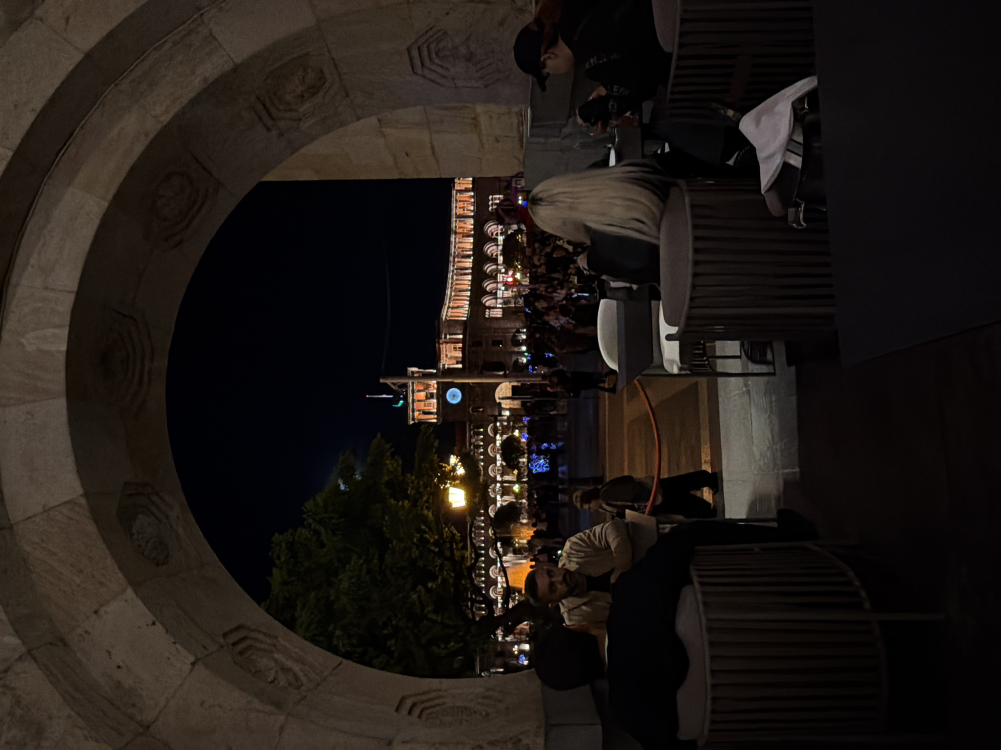 The image shows a nighttime view in Yerevan, Armenia, framed by an arched stone structure. Through the arch, a city square is visible, illuminated by streetlights and decorated with festive lighting. Several people are present in the square, though details are somewhat indistinct due to the distance and lighting conditions. Closer to the viewer, individuals are seated at tables and chairs that appear to be part of an outdoor cafe or restaurant. Some people are engaging with their phones, taking photographs or chatting. One man, sitting at a table with a dark jacket is visible near the bottom-right side of the image. The overall atmosphere suggests a lively social scene in a city center at night.