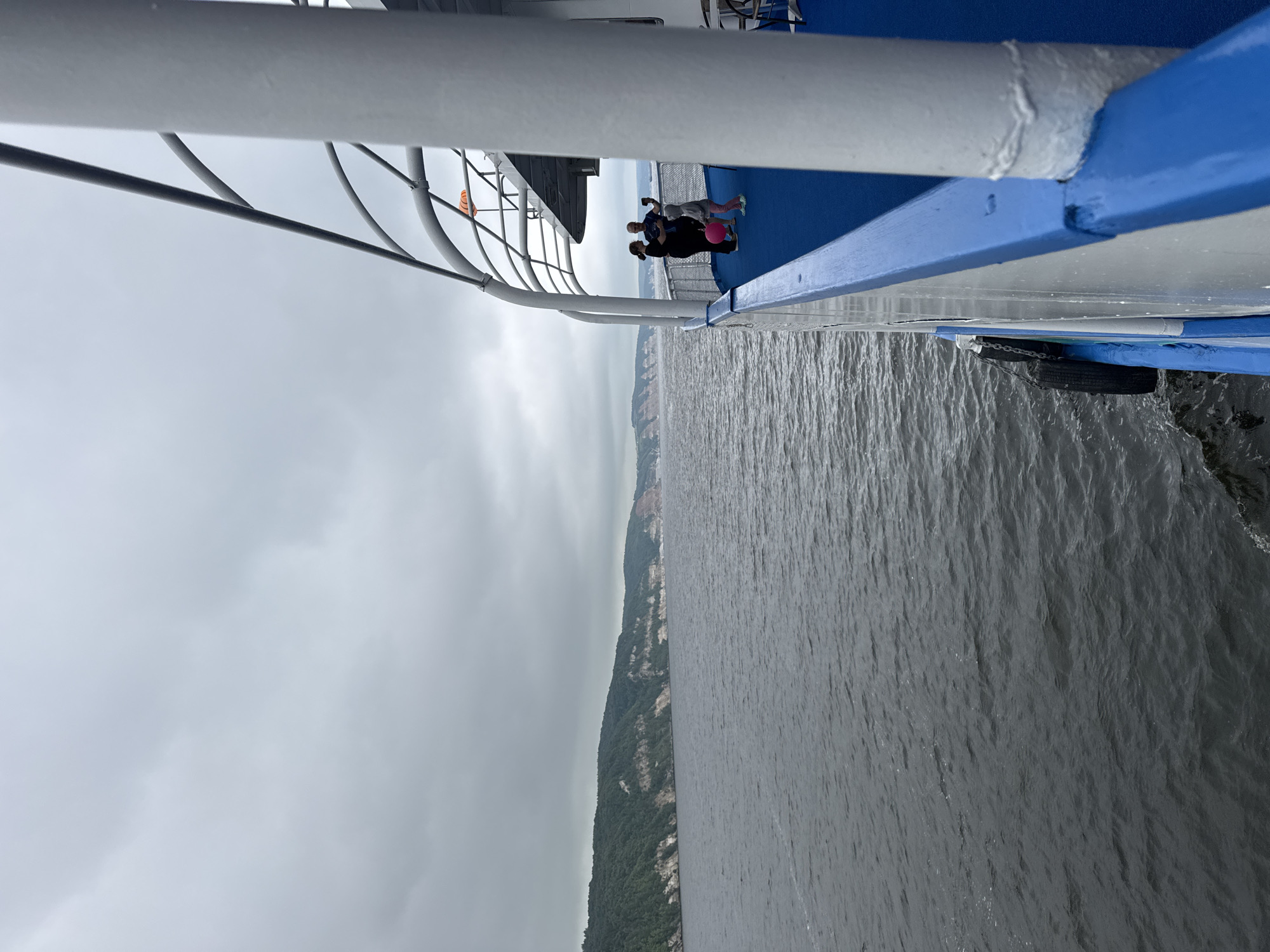 An overcast day at Kamskoye Ustye, Russia, with a view from the deck of a ferry. The dark, choppy water of the Volga River stretches towards a distant, tree-covered bluff. A family of three, a man, woman, and child, are taking a photo on the deck. The woman holds a pink ball. The ferry's blue and white deck railing is prominent in the foreground.