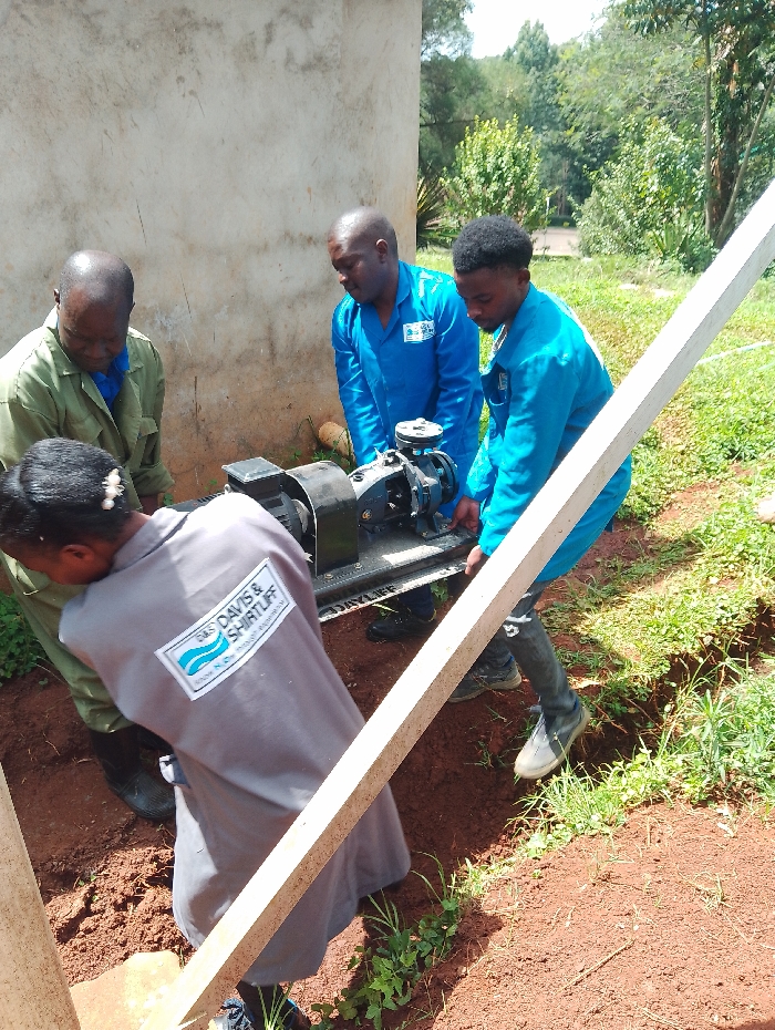 Four individuals are engaged in moving or installing a piece of machinery outdoors in Gitaru, Kenya, under bright daylight conditions.
Three men are actively holding a black pump or motor assembly. The man on the far left wears olive green overalls, while the two men to his right are dressed in blue overalls. One person, seen from behind in the foreground, wears a gray long-sleeved shirt and appears to be assisting with the task. This person's shirt bears a prominent logo on the back, reading "D&S DAVIS & SHIRTLIFF" with the slogan "Water for Life" partially visible beneath it. The machinery itself has the brand name "DAYLIFF" partially legible.
The setting features reddish-brown soil on the ground, with some green grass and scattered vegetation. To the left, a light-colored concrete or stucco wall of a building is visible. The background is filled with lush green trees and foliage, indicative of an outdoor, natural environment. A light-colored, rectangular beam or post diagonally crosses the lower right portion of the frame. The individuals appear focused on their collaborative task.