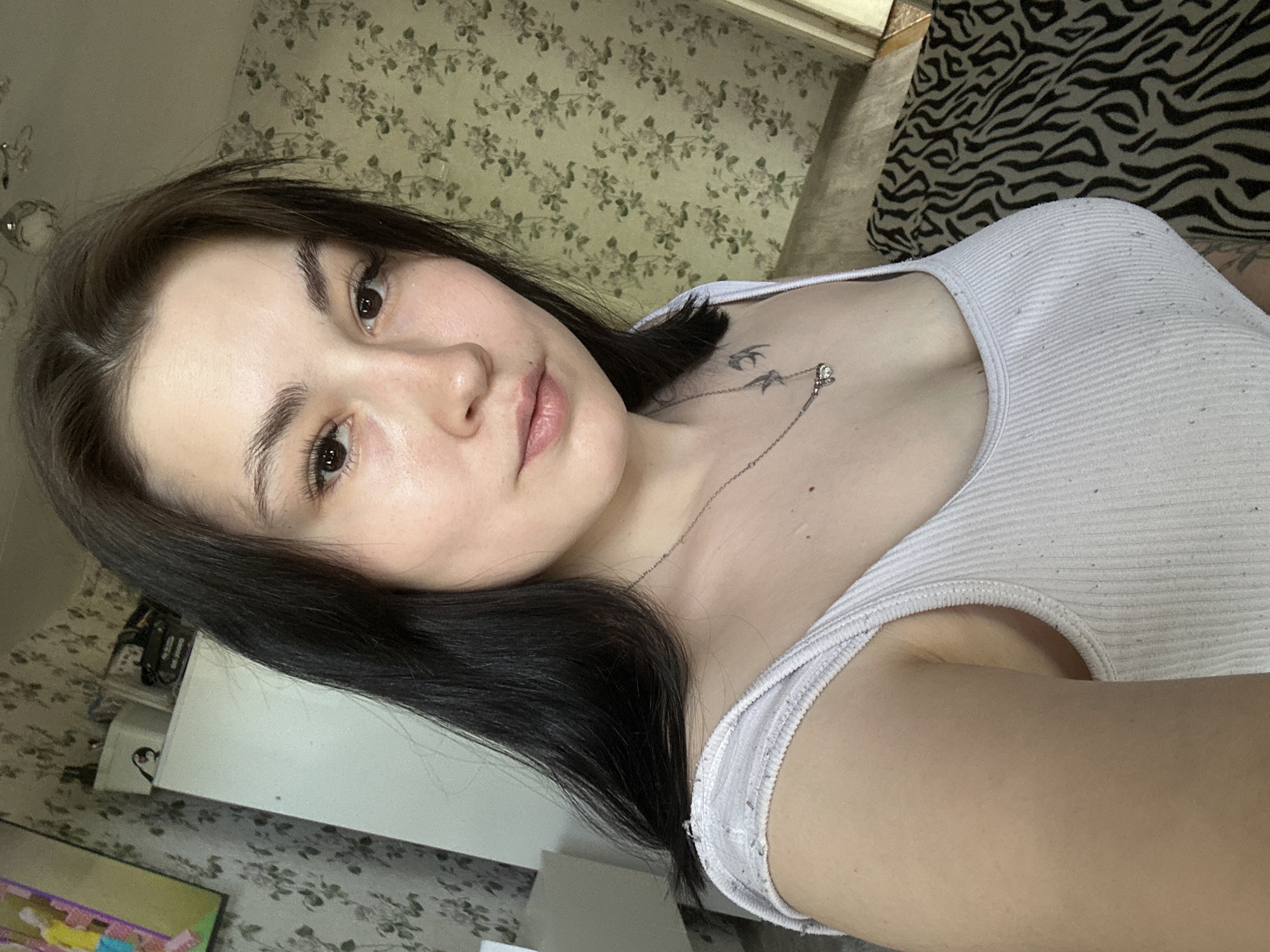 A woman with dark hair and fair skin is depicted in a selfie taken at an oblique angle. She wears a white tank top and a delicate silver necklace with a pendant. A small tattoo is visible on her chest. The background features a floral patterned wallpaper and part of a black and white patterned bedspread. A white cabinet is also visible in the background. Based on the request, the location is specified as Chaikovsky, Russia.