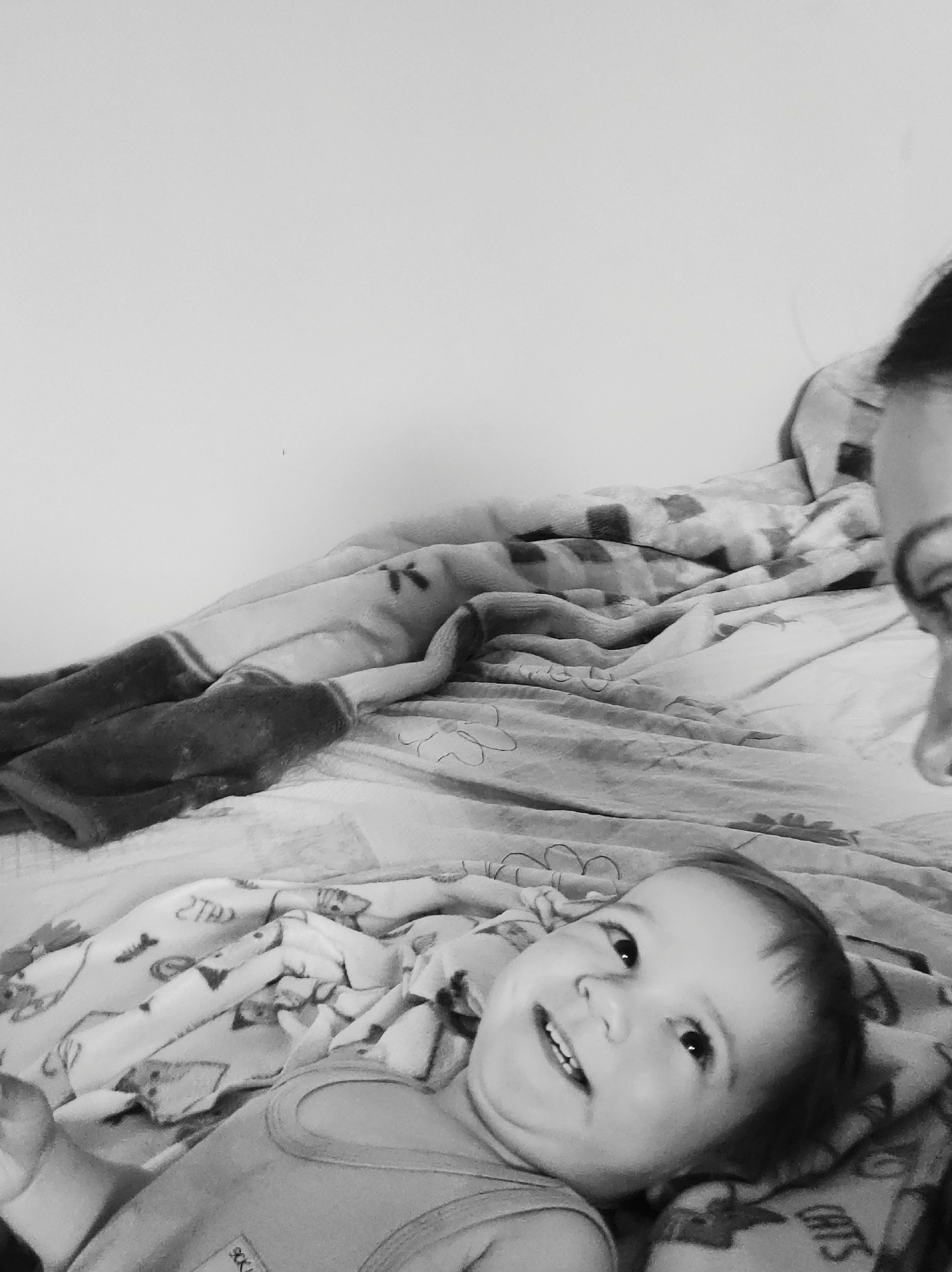 A black and white photograph depicts a young child lying on a bed, with a partially visible adult figure above. The child, in the foreground, is facing upwards with a smile and visible teeth. The child is wearing a tank top. The bed is covered in patterned blankets and a comforter. The adult's face is partially visible, focusing on the eye area. The background features a plain wall. The location is not specified.
