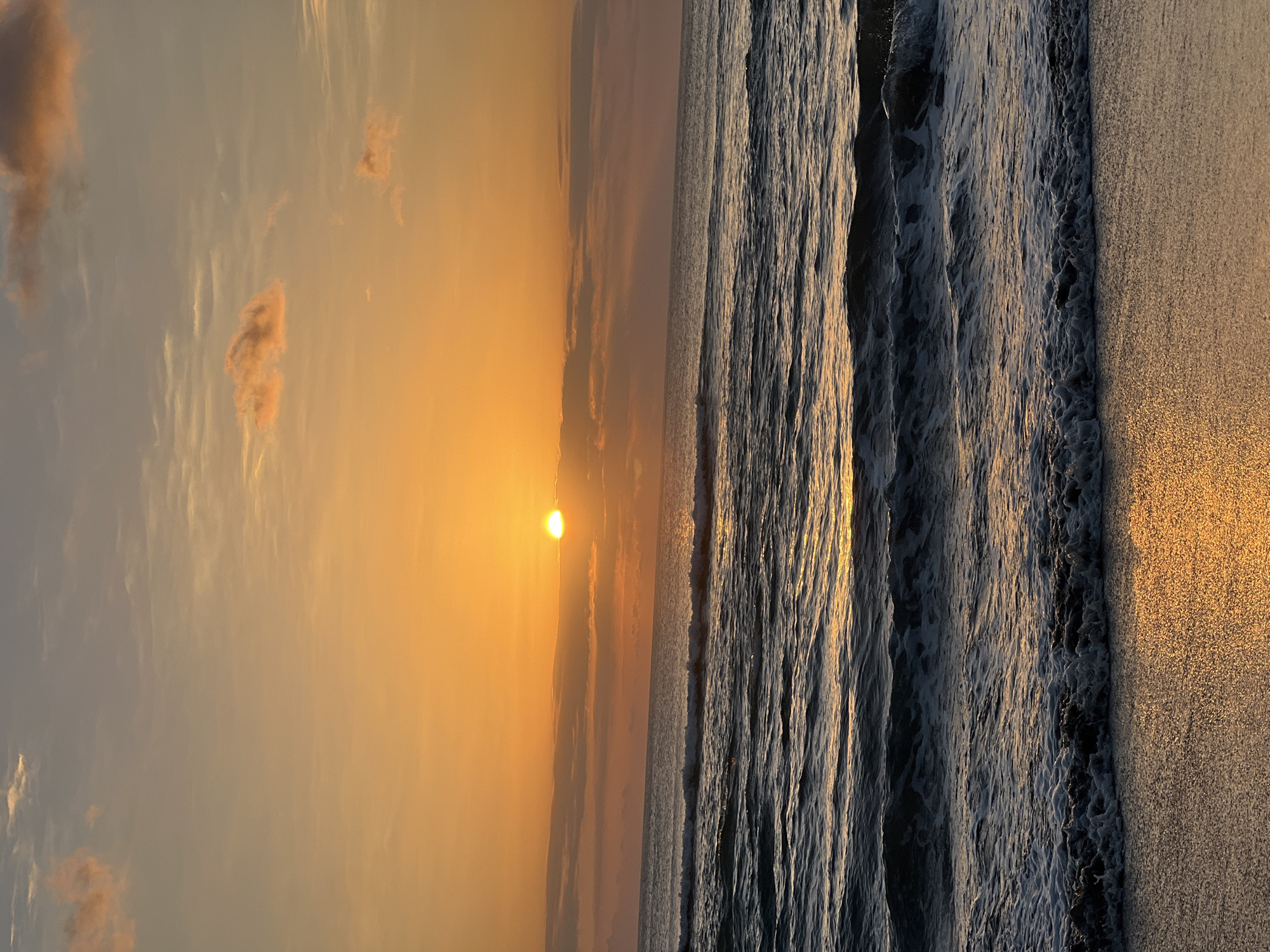 The image depicts a serene ocean sunset at Kuta Beach in Bali, Indonesia. The sun, a bright orange orb, is partially obscured by soft clouds on the horizon, casting a warm glow across the sky and water. Gentle waves roll onto the sandy shore, their crests tinged with golden light. The sand itself reflects the sunset's colors, creating a shimmering pathway. The environment is characterized by the vast expanse of the ocean meeting the tranquil beach under a colorful sky. There are no people visible in the scene.