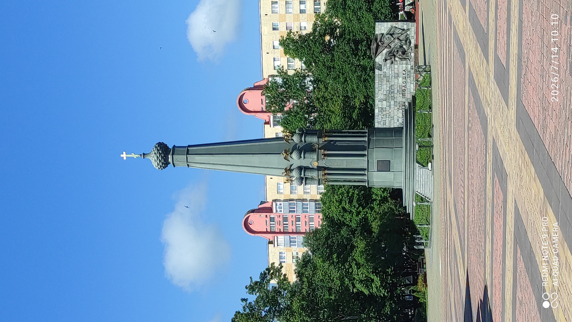 The image depicts a memorial monument in Polatsk, Belarus, on a clear day. The monument is a tall, slender obelisk with a cross at its apex, and it is surrounded by trees and residential buildings in the background. A relief sculpture is visible on a stone wall behind the monument, and the ground in front of it is paved with a geometric pattern of red and yellow bricks. The sky is bright blue with scattered white clouds. There are no people clearly visible in the image, but there is a bench in the background, suggesting a public space. The image includes a watermark in the bottom left corner indicating "REDMI NOTE 9 PRO AI QUAD CAMERA" and a date and time stamp in the bottom right corner reading "2026/7/14 10:10".