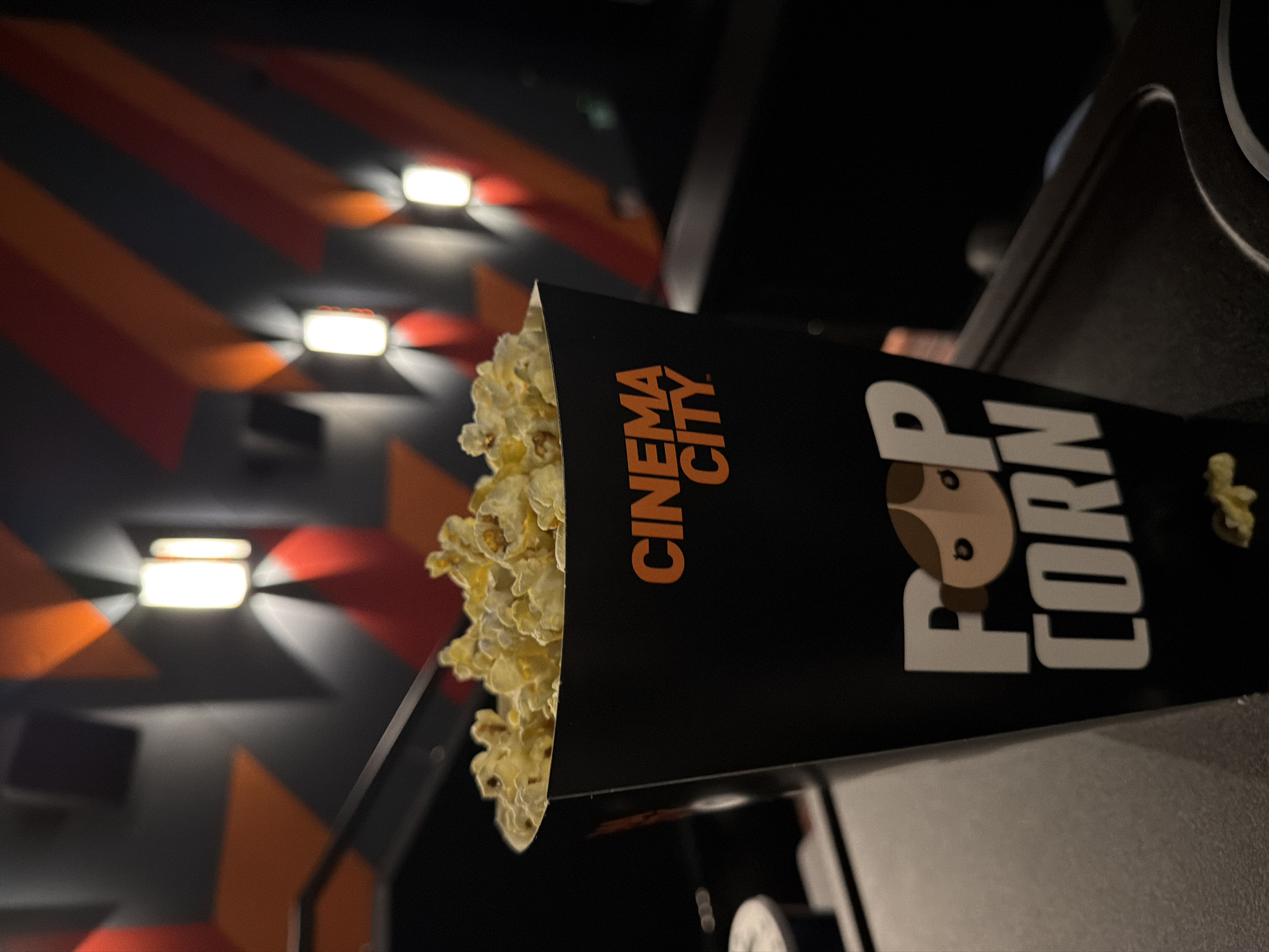 A black popcorn container, overflowing with golden-yellow popcorn, is the central subject of the image, angled slightly to the right. The container features "CINEMA CITY" in orange text on one side and "POP CORN" in white text on the other, with a stylized, light brown dog-like character forming the 'O' in "POP".
The scene is set inside a dimly lit movie theater. In the background, a dark wall or ceiling is visible, adorned with angular, geometric patterns in shades of orange, red, and dark grey. Several bright, rectangular wall lights are mounted on this surface, emitting distinct beams of light that highlight the patterns and contribute to the ambient glow.
A dark, possibly grey, surface, resembling a seat armrest or cup holder, is partially visible in the lower foreground and to the right, suggesting the popcorn container is placed there. The overall lighting is low, typical of a cinema environment before or during a movie screening. No people are visible in the frame. The "Cinema City" branding and the theater setting are consistent with the location context of Sofia, Bulgaria.