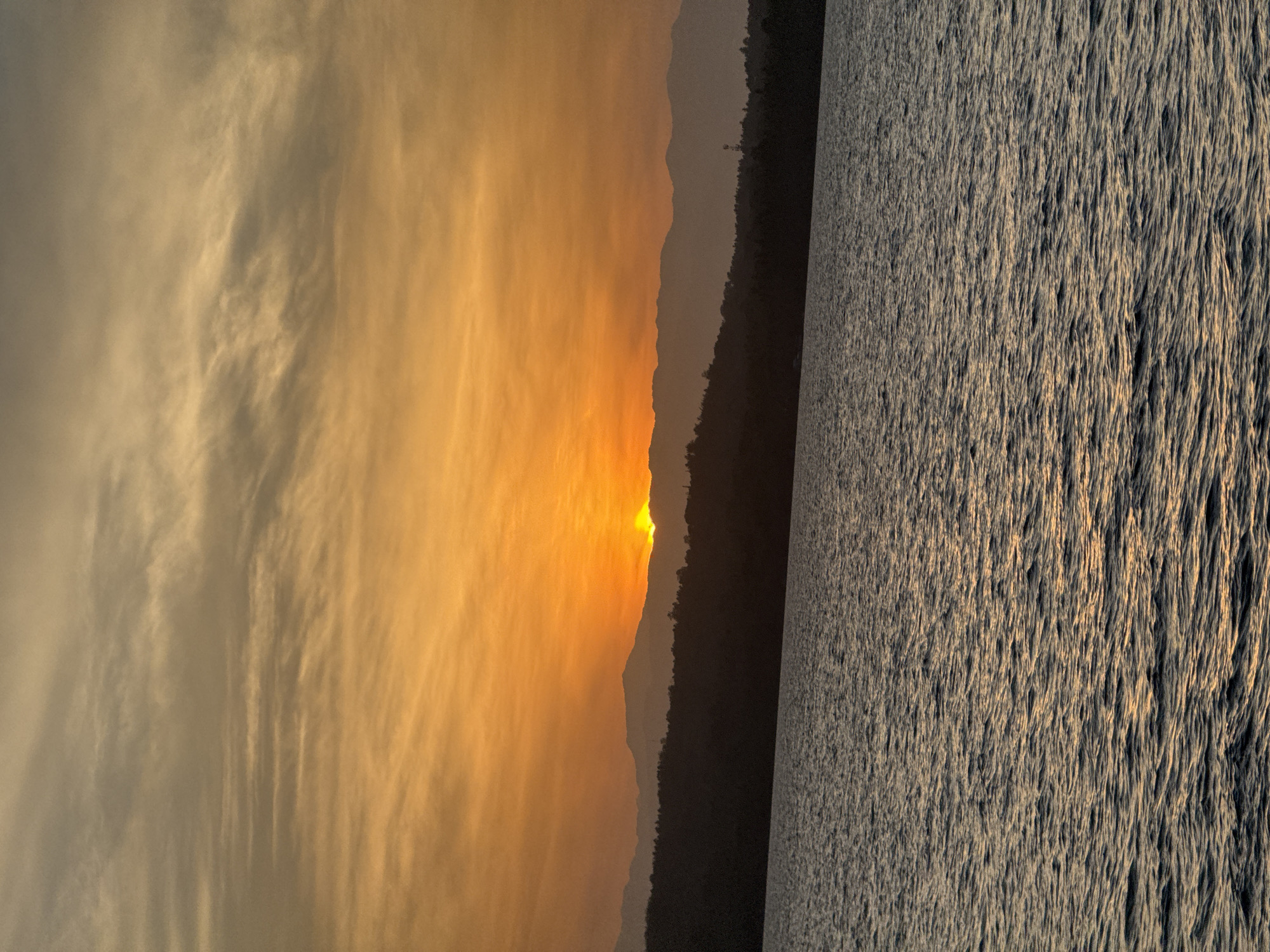 The image shows a sunset over a body of water, likely a lake or ocean, with mountains in the distance. The sky is filled with orange and yellow hues, indicating the sun is near the horizon. The water surface reflects the light and appears textured with small waves. The mountains form a dark silhouette against the bright sky. The location is not possible to determine solely based on the image without additional information.