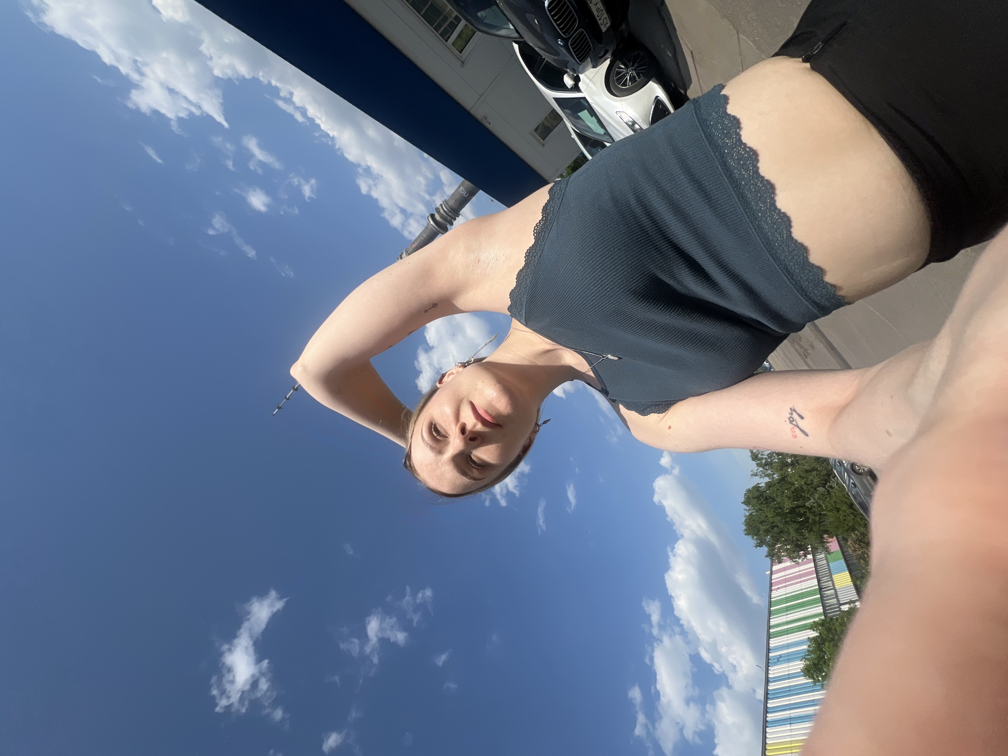 A young woman in Khimki, Russia takes a selfie. She wears a dark blue lace-trimmed tank top and black shorts. A car and a building with a colorful facade are visible in the background. The sky is blue with scattered clouds.