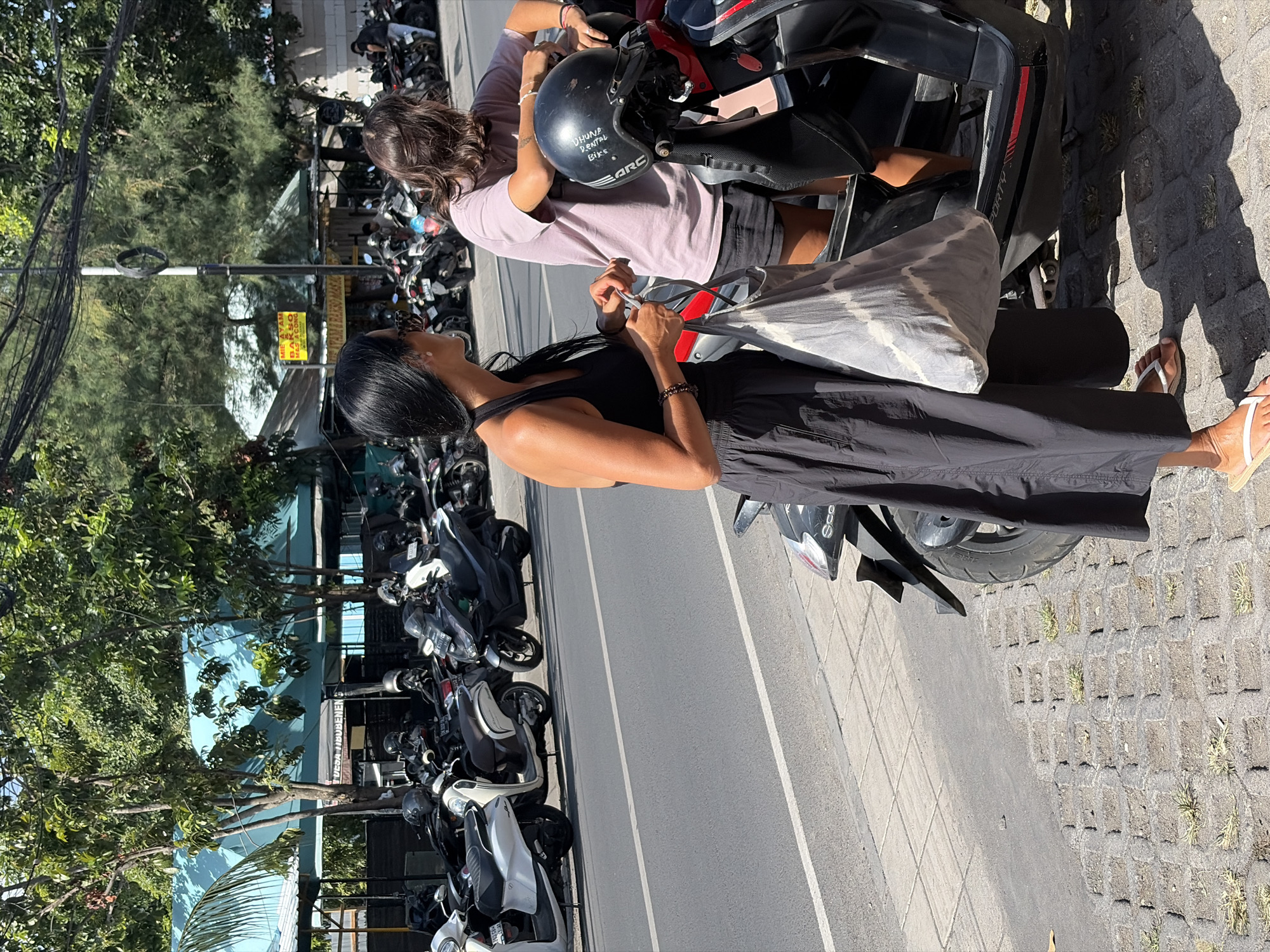 A medium shot captures two women standing next to a parked motorcycle on a street in North Kuta, Indonesia. The woman on the left, with long dark hair, wears a black tank top and a long black skirt, carrying a grey and white tie-dye tote bag. The woman on the right, with long brown hair, is wearing a light purple t-shirt and dark shorts. A black motorcycle helmet with white writing is attached to the handlebars of the motorcycle. In the background, numerous other motorcycles are parked, and lush green trees line the street. A blue awning stretches across a building in the far background. The ground is paved with textured concrete squares. The lighting suggests it is daytime.