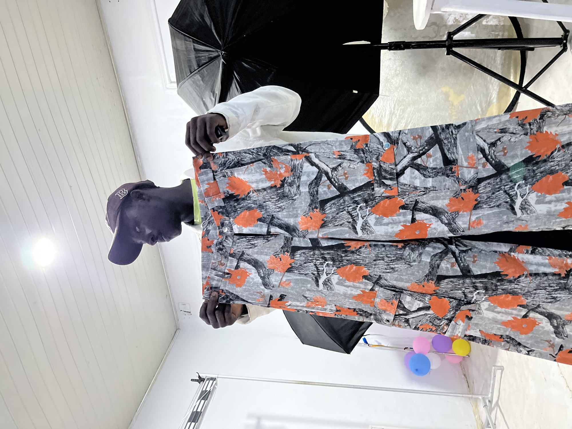 Inside a brightly lit photography studio in the city of Unknown, Unknown, a male individual of dark complexion wears a brown baseball cap with a subtle "BB" logo and a white long-sleeved shirt over a neon green undergarment. He stands holding up a pair of cargo pants featuring a unique camouflage pattern of grey and black tree branches interspersed with prominent bright orange maple leaves. His gaze is directed towards the upper right, off-camera. In the background, a white slatted ceiling with a central illuminated light fixture is visible. To the left, a white metal clothing rack stands near a cluster of colorful balloons (blue, pink, purple, yellow). To the right, a large black photographic umbrella softbox is set up on a tripod, partially obscuring another white surface or reflector behind it. The walls are plain white, and the floor is a light, possibly tiled surface. A segment of a white cylindrical stool or table is visible on the far right.