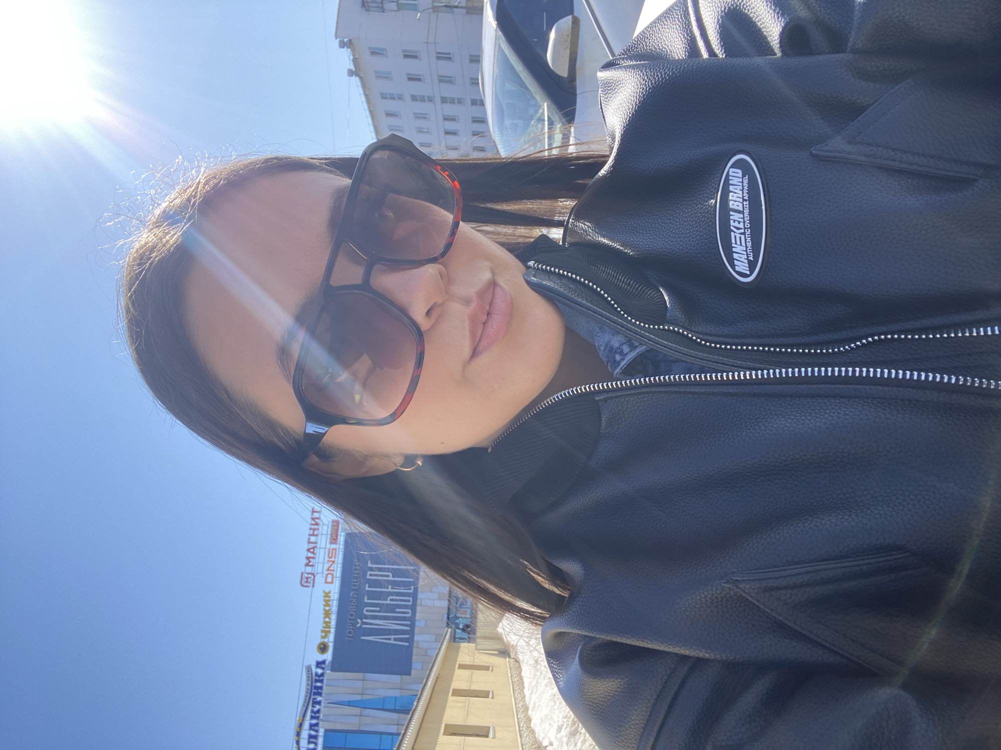 A woman poses for a selfie in Chaikovsky, Russia, on a sunny day. She is wearing dark sunglasses and a black leather jacket with a "MANEKEN BRAND" patch. In the background, a building with signs for "МАГНИТ", "DNS", and "АЙСБЕРГ" is visible.