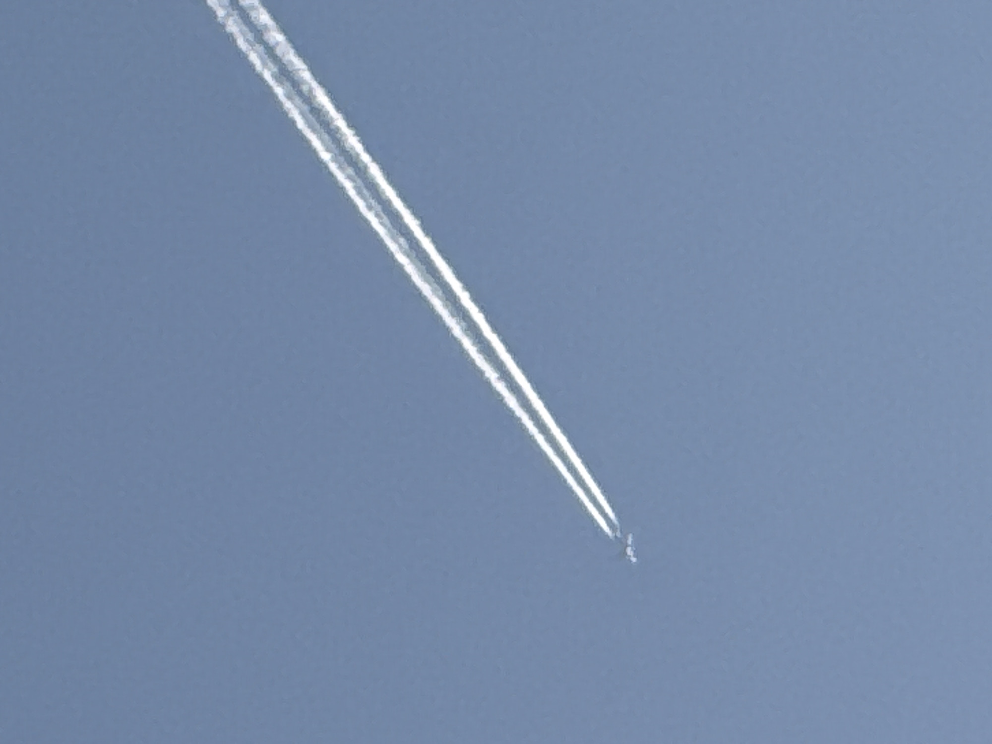 A daytime, aerial view captures a contrail emitted by a high-altitude aircraft. The contrail is white and trails from the upper-left toward the lower-right of the frame, terminating where the aircraft is visible, mid-sky. The background is a clear, light blue sky. The location is unspecified and cannot be determined from the image.