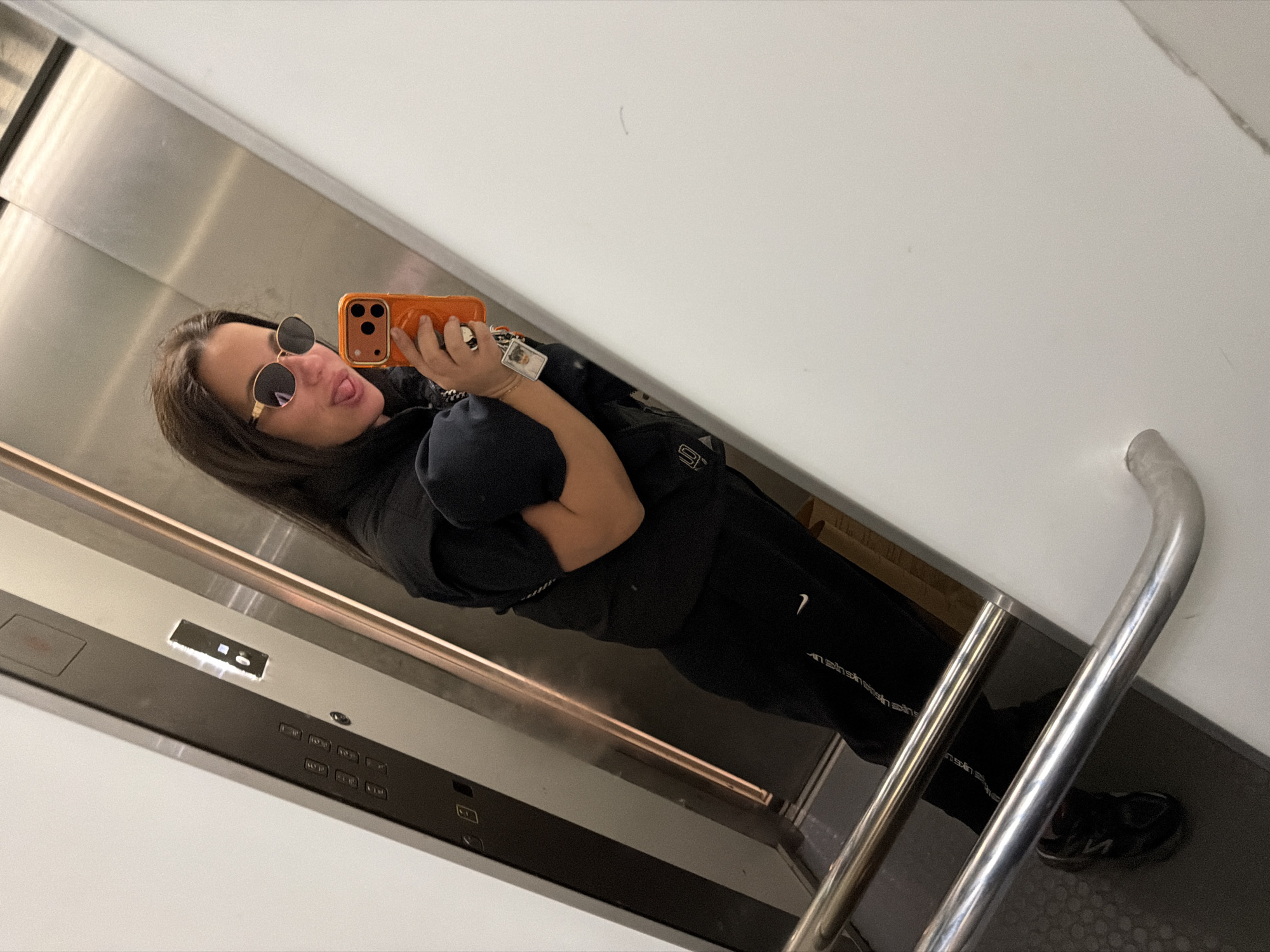 A woman in an elevator takes a selfie with an orange phone. She is wearing sunglasses, a black puffer vest, and a black tracksuit with a white Nike logo. The elevator has a stainless steel interior, and a control panel is visible. A silver handrail is on the right. The photo's location is unidentifiable as Burgas, Bulgaria.