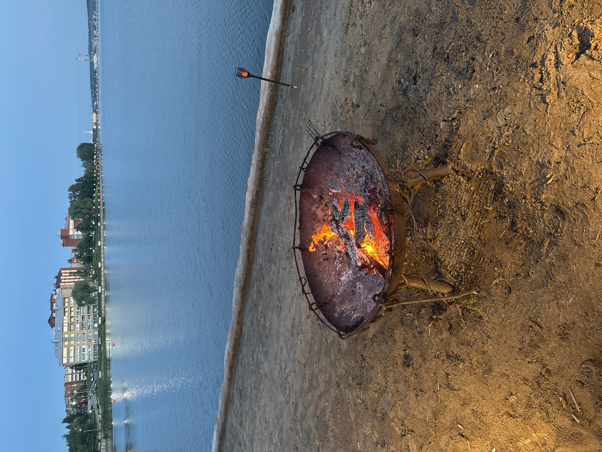 This image captures a twilight or nighttime scene along the Votkinsk Pond/Reservoir in Votkinsk, Russia. In the immediate foreground, a dark, rustic metal fire pit with three legs rests on sandy ground, containing glowing embers and actively burning wood logs that cast an orange-yellow light. To the right of the fire pit, a dark-colored torch on a stake is embedded in the sand, emitting a consistent orange, flame-like light. A light-colored, elevated bank, possibly concrete or compacted earth, separates the sandy area from the main body of water. The water itself is dark blue and calm, reflecting ambient light. Two small, bright orange buoys are visible floating on the water in the left-mid distance. Along the far shoreline, the city of Votkinsk is visible. A line of streetlights illuminates a promenade or pathway, behind which multiple multi-story buildings are present. A prominent, light-colored apartment building with illuminated windows is situated in the left-center background. Various other structures, including buildings with red roofs and more distant industrial elements such as two tall chimneys, are visible stretching across the horizon. The sky is a deep, clear twilight blue.