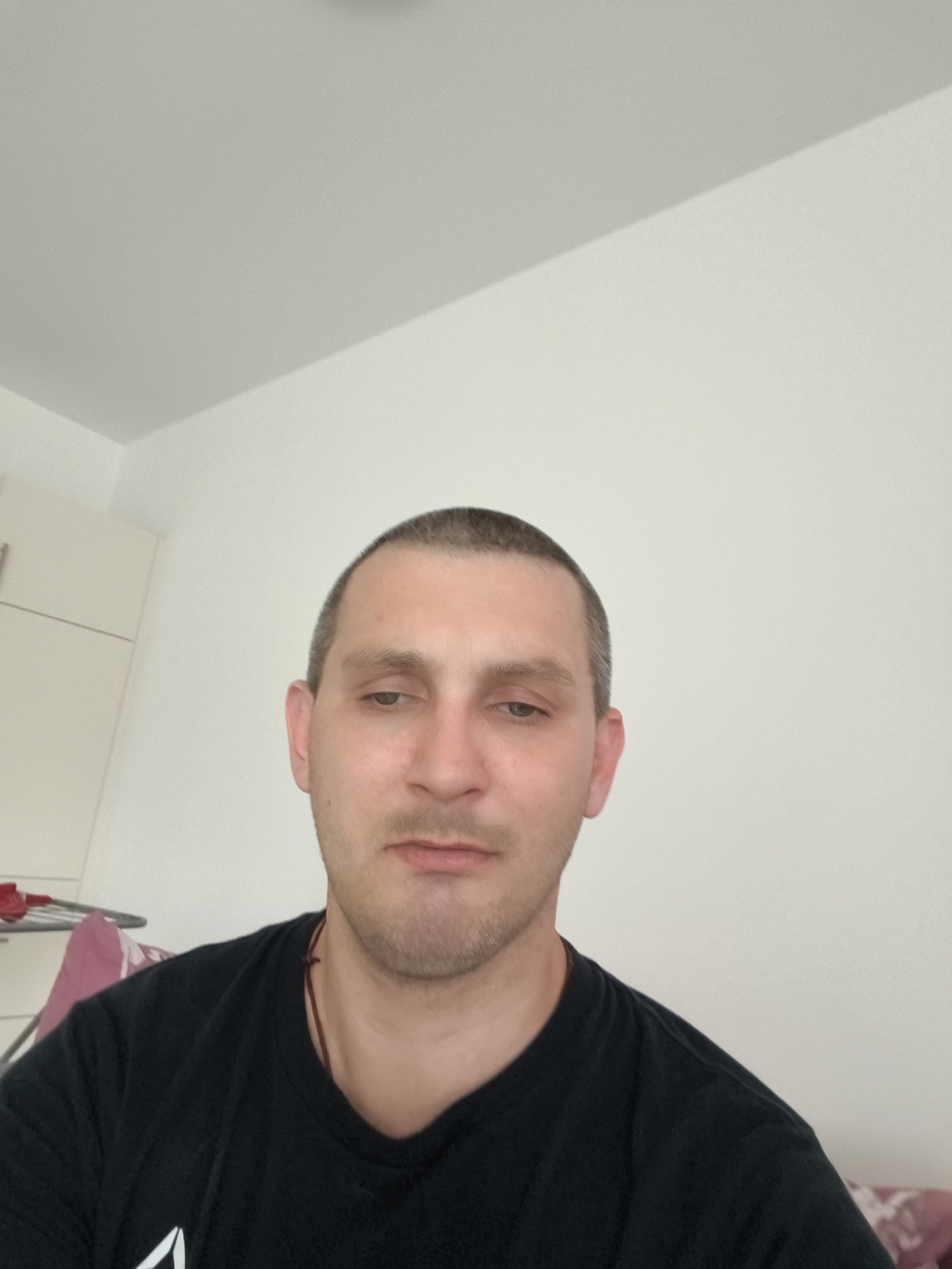 An overhead, close-up, medium shot shows a man in his late 20s looking downwards. The man has a shaved head and is wearing a black t-shirt. He has stubble on his face. The background appears to be a room with a white wall and part of a cabinet. The lighting suggests it is daytime.