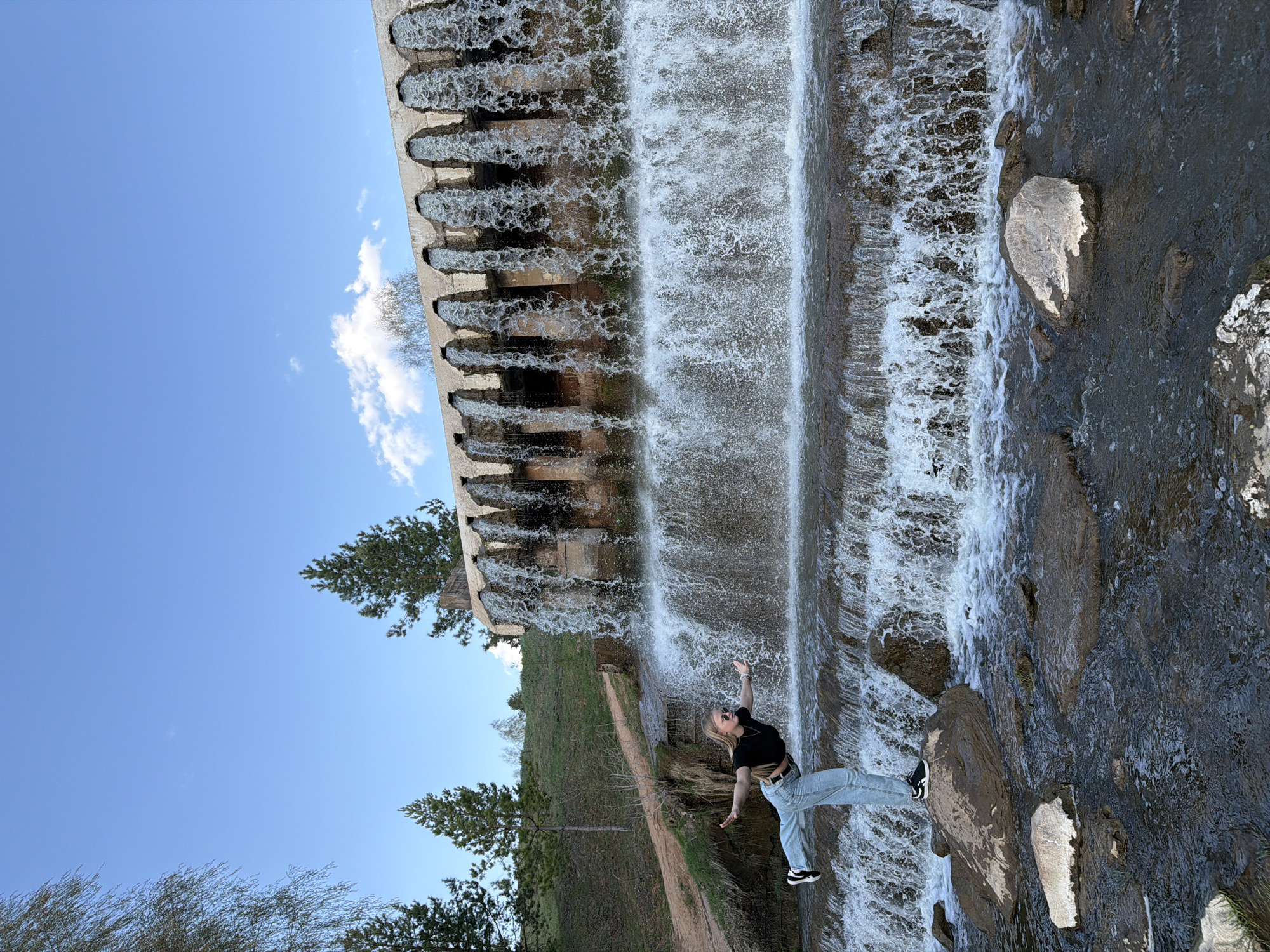 A medium shot shows a woman standing in front of a tiered waterfall at Kamennoye, Russia. The woman has her arms outstretched. She is wearing blue jeans, a black shirt, and black and white tennis shoes. The waterfall is to the left. A gray stone structure releases water from the top of the waterfall. The sky is blue with a few white clouds. Trees are located at the base of the waterfall.