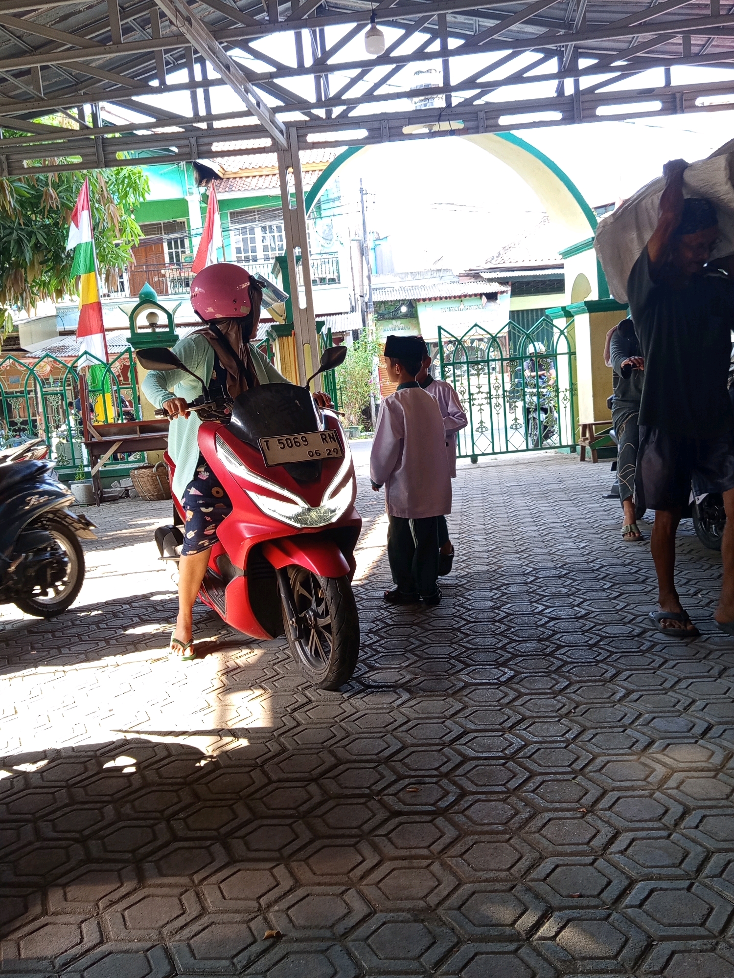The image depicts an outdoor scene in Karawang, Indonesia, likely in the vicinity of a mosque or religious institution, given the presence of green gates and a minaret visible in the background. In the foreground, a woman wearing a light blue hijab, a blue patterned dress, and a pink helmet is seated on a red scooter. She is facing towards the viewer's left. To the left of the scooter, another black scooter is partially visible.
In the middle ground, two young boys in white religious attire and black skullcaps are standing and looking towards the left. Behind them, a person is carrying a large white sack over their shoulder. Further back, a building with green and yellow accents and arched windows is visible. Indonesian flags, one red and white, and another with yellow and green stripes, are also present.
The ground is paved with interlocking hexagonal bricks. Sunlight casts strong shadows across the paving stones, indicating it is daytime. The overall impression is of a community gathering or a scene outside a religious site on a typical day in Karawang.