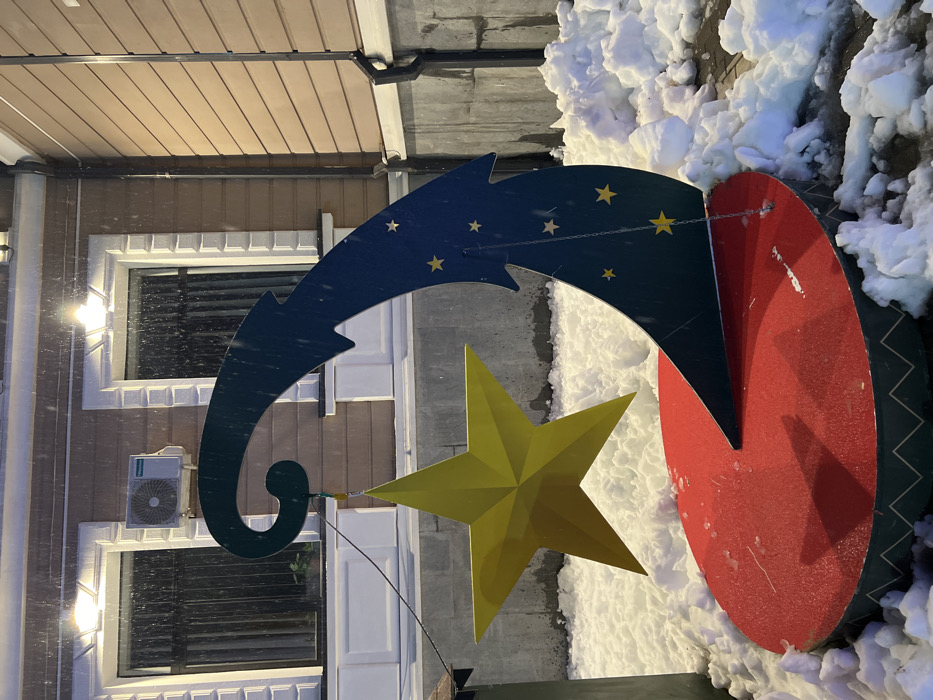 This image depicts an outdoor decorative display in Terjola, Georgia, during what appears to be snowfall at night. The primary subject is a holiday-themed installation consisting of a large, three-dimensional yellow five-pointed star suspended from a dark blue, upward-curving stylized wave or crescent moon structure. This dark blue element is adorned with multiple smaller yellow stars and is anchored to a red circular base. The base features a dark green rim with a white zigzag pattern. The ground surrounding the installation is covered with irregular mounds of white snow.
In the background, a light brown building with horizontal siding is visible. Two symmetrically placed windows with ornate white sills and frames are present, each equipped with dark vertical blinds or curtains internally. An external air conditioning unit, identified as Hisense, is mounted on the wall between the two windows. Two wall-mounted floodlights illuminate the scene, with visible precipitation in the air. A dark vertical downspout runs along the right side of the building. The lower portion of the building features a grey, textured concrete foundation. No individuals are visible in the scene.