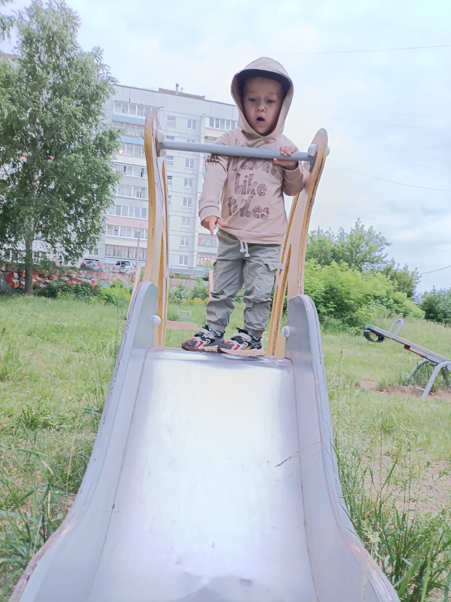 A young boy, appearing to be around 3-4 years old, stands at the top of a metal slide on a cloudy day. He is wearing a beige hooded sweatshirt with the words "I don't like rules" printed on it in a darker shade, and olive green cargo pants. His shoes are a mix of black, white, and red. The slide is part of a playground structure with wooden supports. In the background, a multi-story apartment building with light-colored facades and numerous windows is visible. To the left of the building, a tall tree with green foliage stands. The foreground and midground are covered in green grass. A seesaw is visible in the right background. The overall setting is an urban playground area in Chaikovsky, Russia.