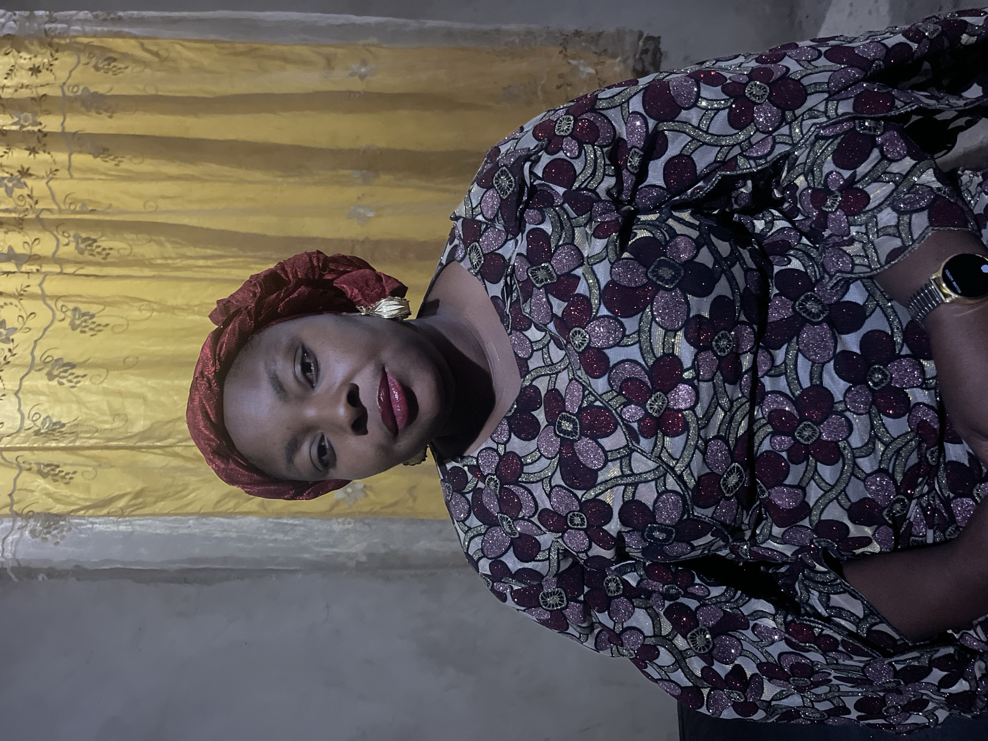 A medium shot captures a woman from the chest up, seated in an indoor setting. She is wearing a red headscarf and a patterned dress with floral motifs in shades of burgundy, purple, and gold against a cream background. Large gold earrings adorn her ears. Her gaze is directed forward, and her lips are painted with red lipstick. The background features a wall on the left and a yellow curtain with embroidered floral designs on the right. A part of a black chair is visible on the lower left. A gold watch is visible on her left wrist. The lighting appears to be artificial, casting subtle shadows. The overall scene suggests a formal or semi-formal occasion.