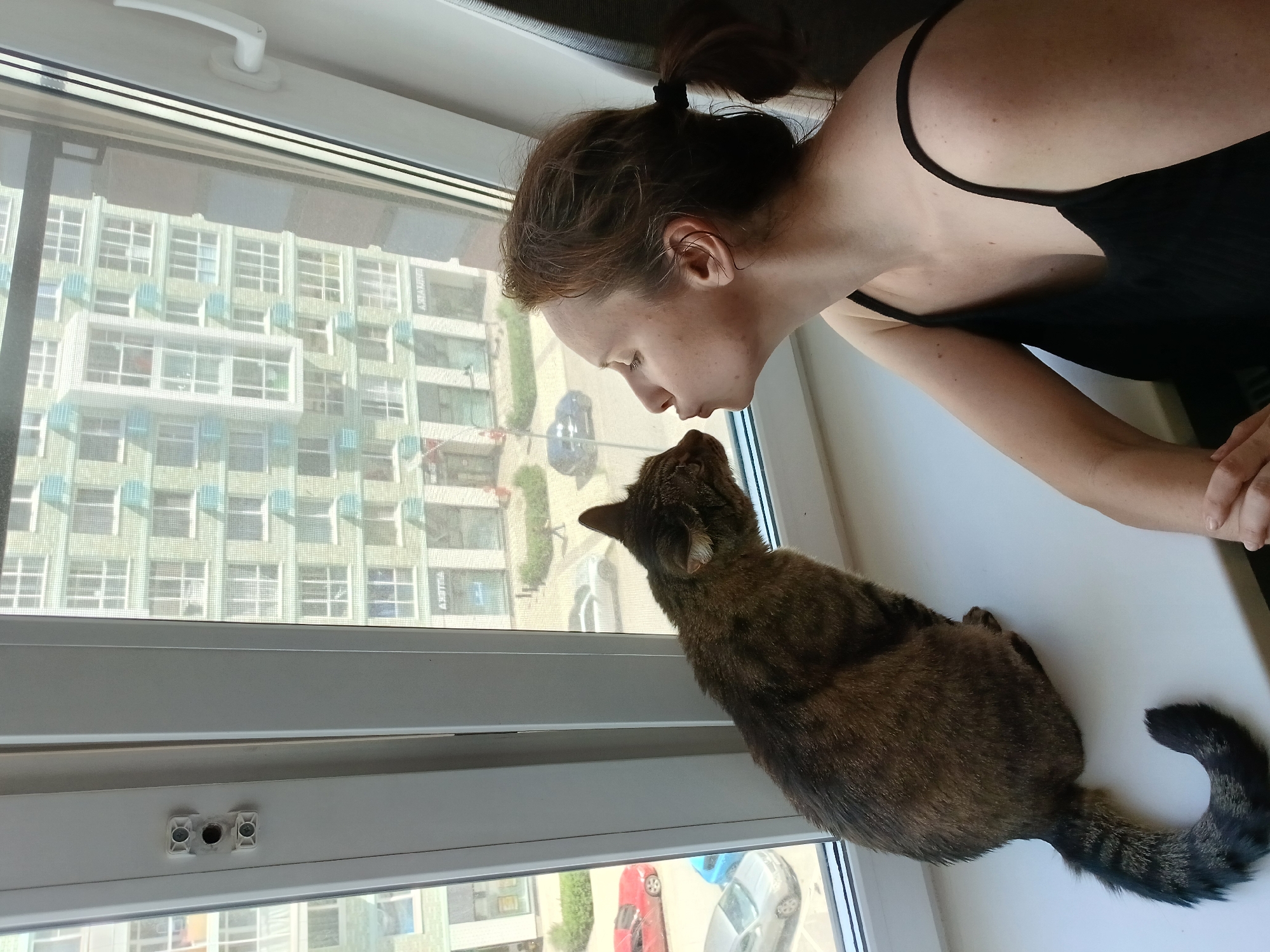 In Yekaterinburg, Russia, a woman with brown hair tied in a ponytail, wearing a black tank top, sits on a white windowsill. She is leaning towards a tabby cat, also on the windowsill, and is puckering her lips as if to kiss it. The cat is facing the woman and appears to be looking at her. They are positioned in front of a large window that looks out onto a city street. Across the street, a multi-story building with a light green facade and numerous windows is visible. Cars are parked on the street below, and a person can be seen walking on the sidewalk. The interior of the room appears to be modern, with a white window frame and trim.