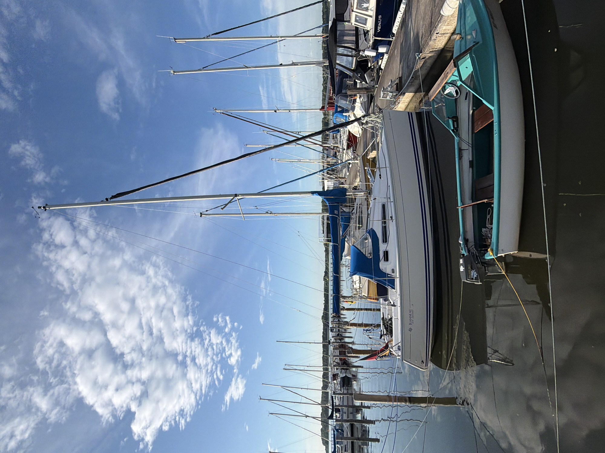 This media file shows a marina with numerous sailboats docked in Ribnitz-Damgarten, Germany. The scene is captured on a clear day with scattered white clouds in a bright blue sky, suggesting it is likely daytime, possibly late morning or afternoon. The water is calm, reflecting the boats and the sky, indicating a peaceful atmosphere.
The primary subjects are the sailboats, with their masts tall and prominent against the sky. One sailboat in the foreground has a white hull with blue stripes and a dark-colored jib. Another teal-colored boat is partially visible. Several other sailboats are docked alongside a concrete pier, lined up neatly.
There are no people visible in the frame. The activity is static, with the boats at rest in the harbor. Notable details include the rigging of the sailboats, the concrete dock, and the reflections in the water. One of the boats has the text "VENUS" and "JEANNEAU" visible on its hull, along with some smaller print that is not clearly legible. The overall impression is one of a serene maritime setting.
