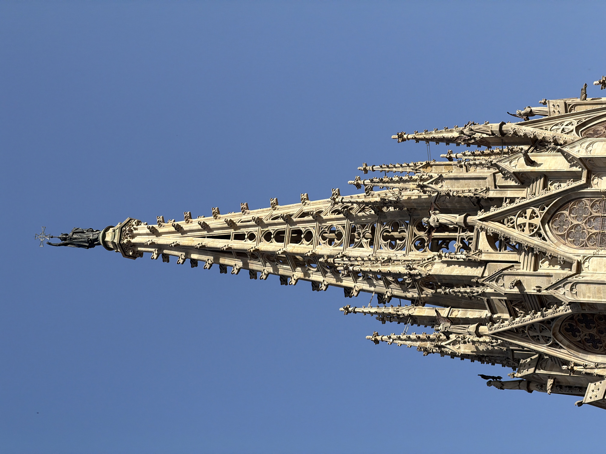 The image shows the spire of the Barcelona Cathedral (Catedral de Barcelona), located in the Gothic Quarter of Barcelona, Spain. The spire is intricately carved stone with multiple spires and statues. At the very top, a dark statue of a figure holding a cross is visible against a clear blue sky. Gargoyles jut out from the sides of the spire. The stone is weathered and appears to be a light beige color. Sunlight casts shadows on the detailed carvings.