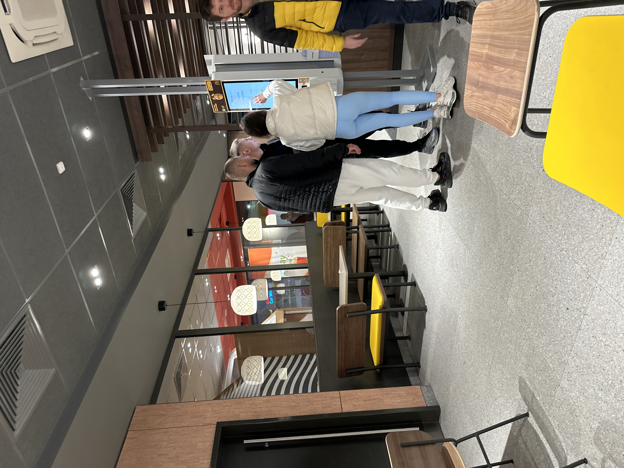 A medium, indoor shot shows four people standing in front of a self-ordering kiosk in what appears to be a McDonald's restaurant in Plovdiv, Bulgaria. The restaurant is decorated with warm wood tones and modern lighting fixtures.
Three individuals are actively engaged with the kiosk, while a fourth person stands slightly to the side. The person closest to the camera wears a black quilted jacket and white pants. The next person has on a white puffer jacket with blue leggings. The third person has on a yellow jacket.
To the right, there are several tables with brown tops and chairs, some with yellow seats. The floor is light gray. A large window with decorative pendant lights illuminates the area. A black doorway is in the bottom left corner.