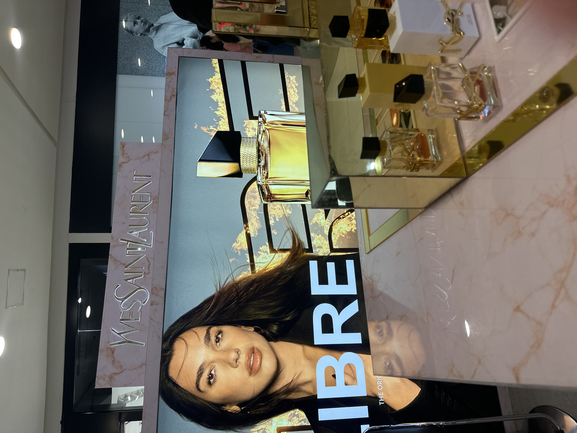An Yves Saint Laurent advertisement is displayed on a screen in a retail store. The advertisement features a portrait of a woman, a bottle of perfume, and the text "LIBRE". In the foreground, several bottles of Yves Saint Laurent perfume are arranged on a marble countertop. The store appears to be located in Davao City, Philippines, as indicated by a small flag that is partially visible in the bottom right corner of the image.