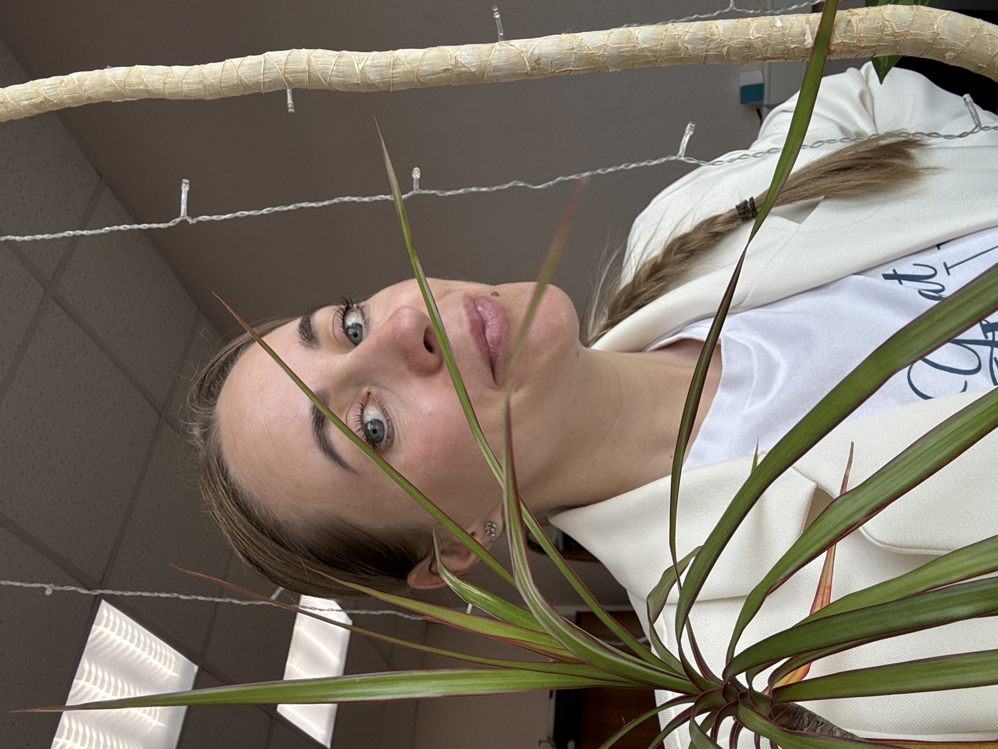 A woman with fair skin, blue eyes, and light brown hair pulled back into a braid, is viewed from below. She is wearing a white t-shirt and a white blazer. Her face is framed by long, spiky green and red leaves. Behind her, there is a horizontal, roughly textured beige branch, adorned with small white fairy lights. The background shows a tiled ceiling and wall, partially illuminated by a window with patterned light filtering through. While the location is given as Chaikovsky, Russia, there is no visual information within the image to definitively confirm this.